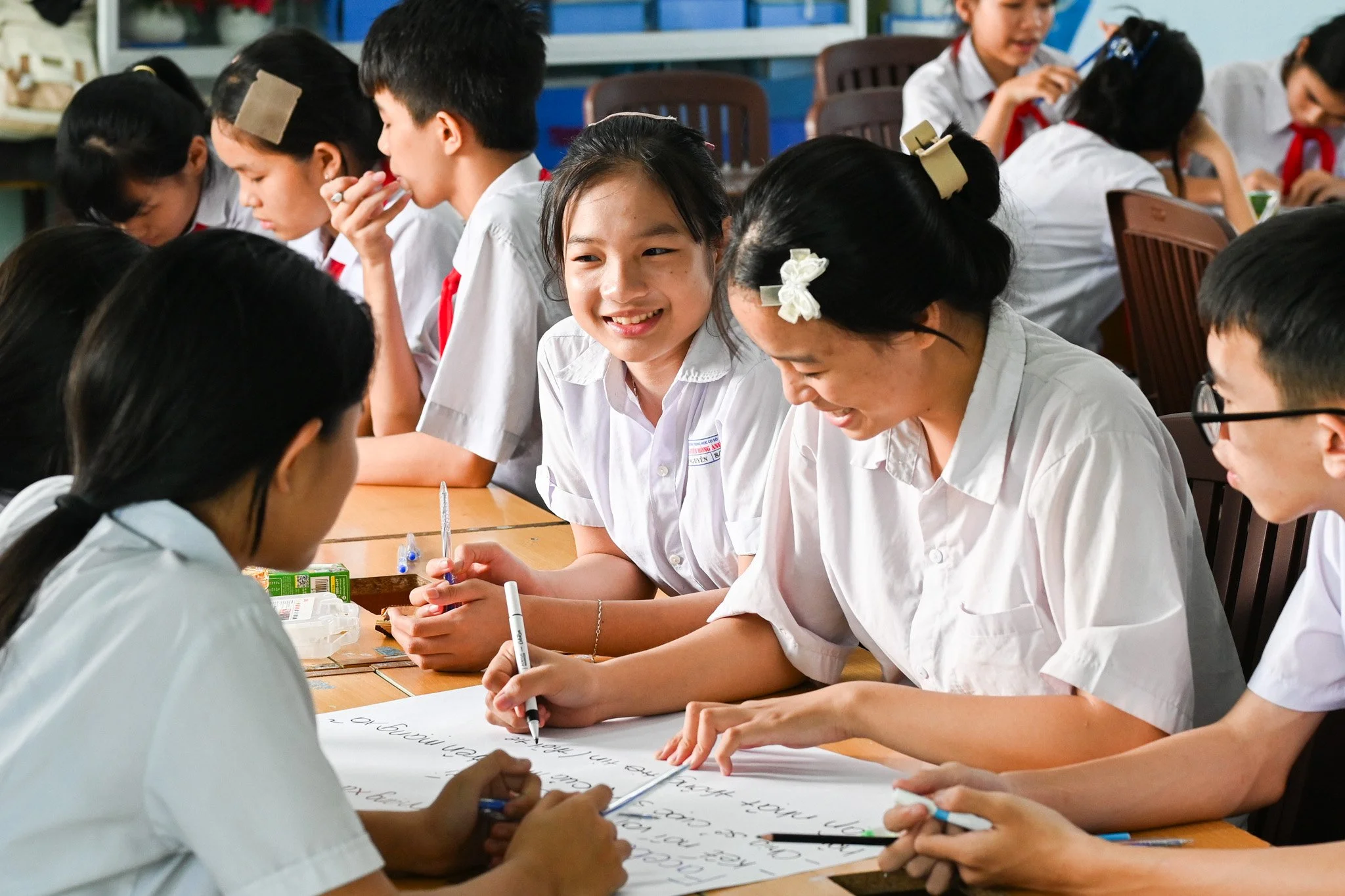 A group of students sitting at a table in a classroom, working on a project or writing on a large sheet of paper, with some students smiling and engaging in discussions.