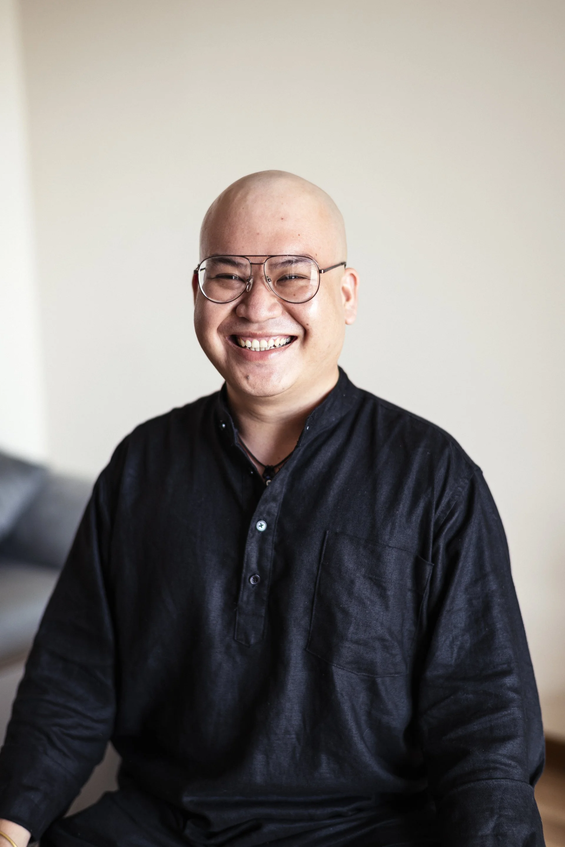 A smiling man with glasses and a shaved head wearing a black button-up shirt, sitting indoors against a plain light-colored wall.