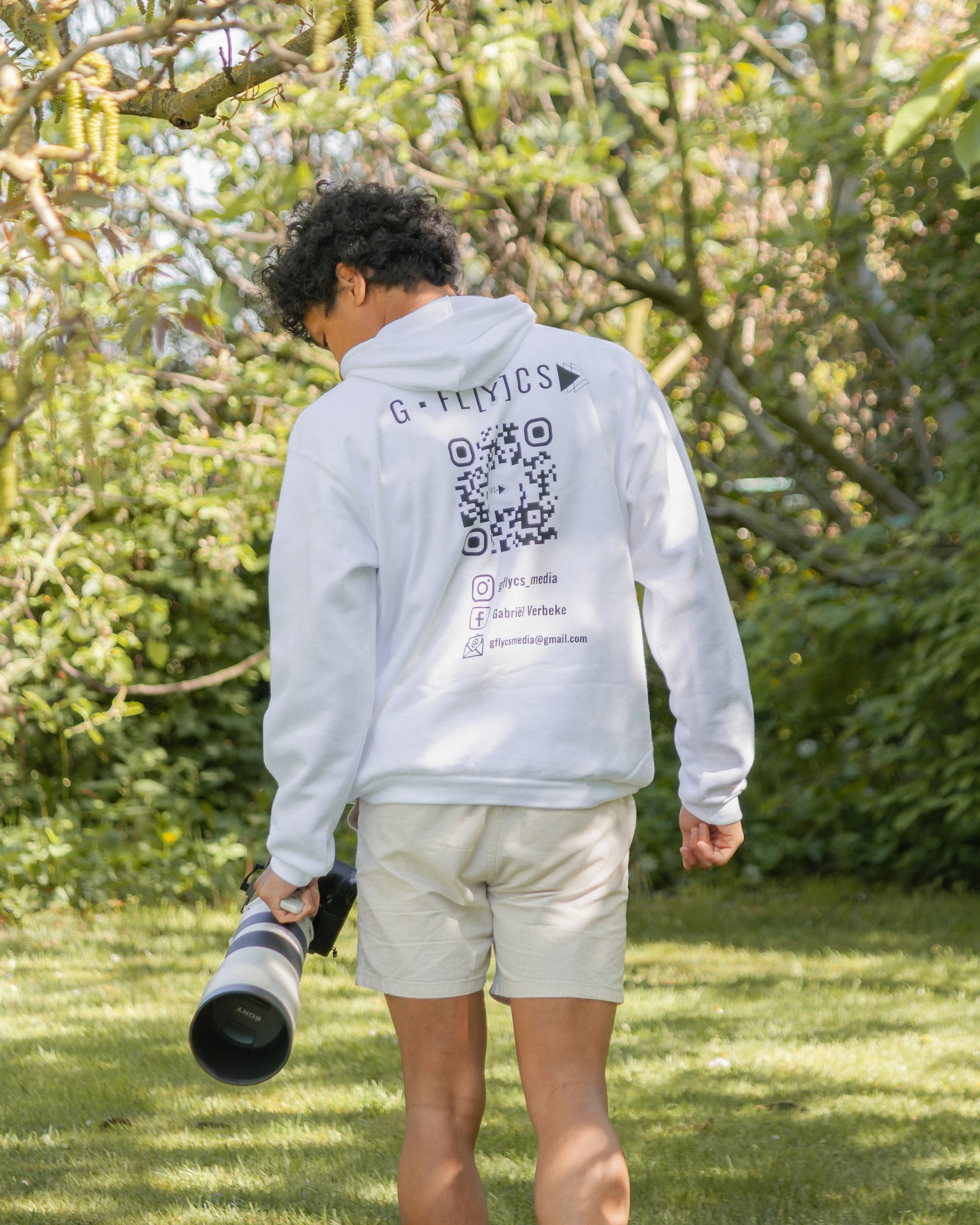 Person walking in a park with trees, carrying a camera with a large lens, wearing a white hoodie with social media logos and a QR code on the back.