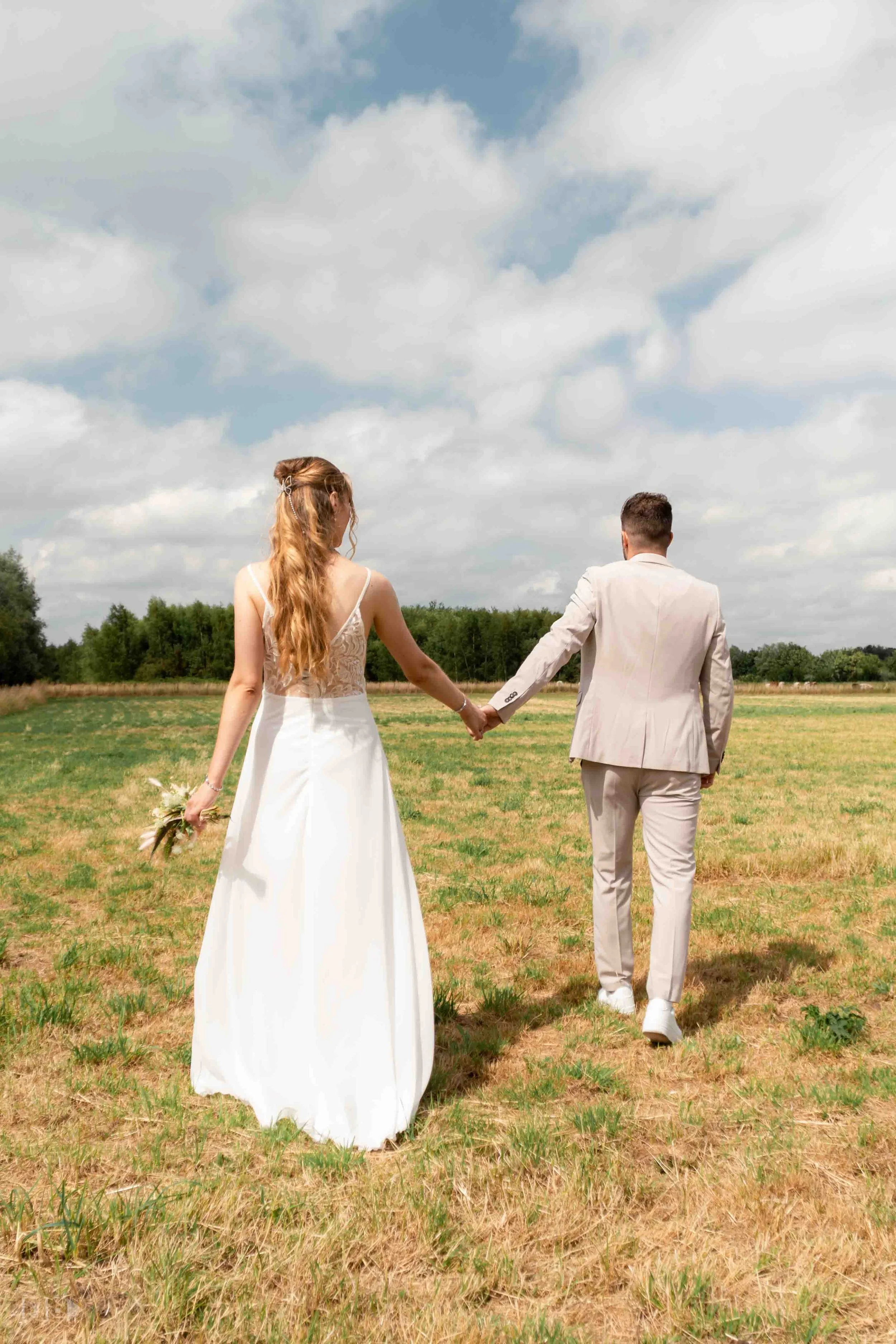 A bride and groom walking hand in hand in an open field on a partly cloudy day.