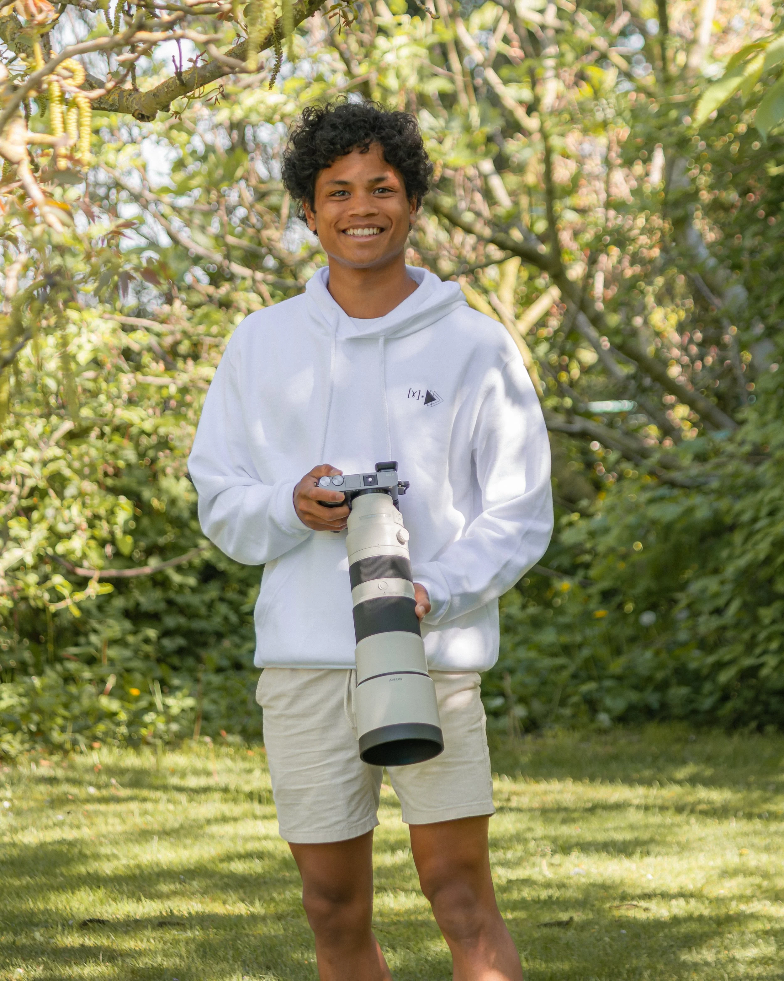 A young man with curly black hair smiling in a garden, holding a professional camera with a long lens, wearing a white hoodie and beige shorts.