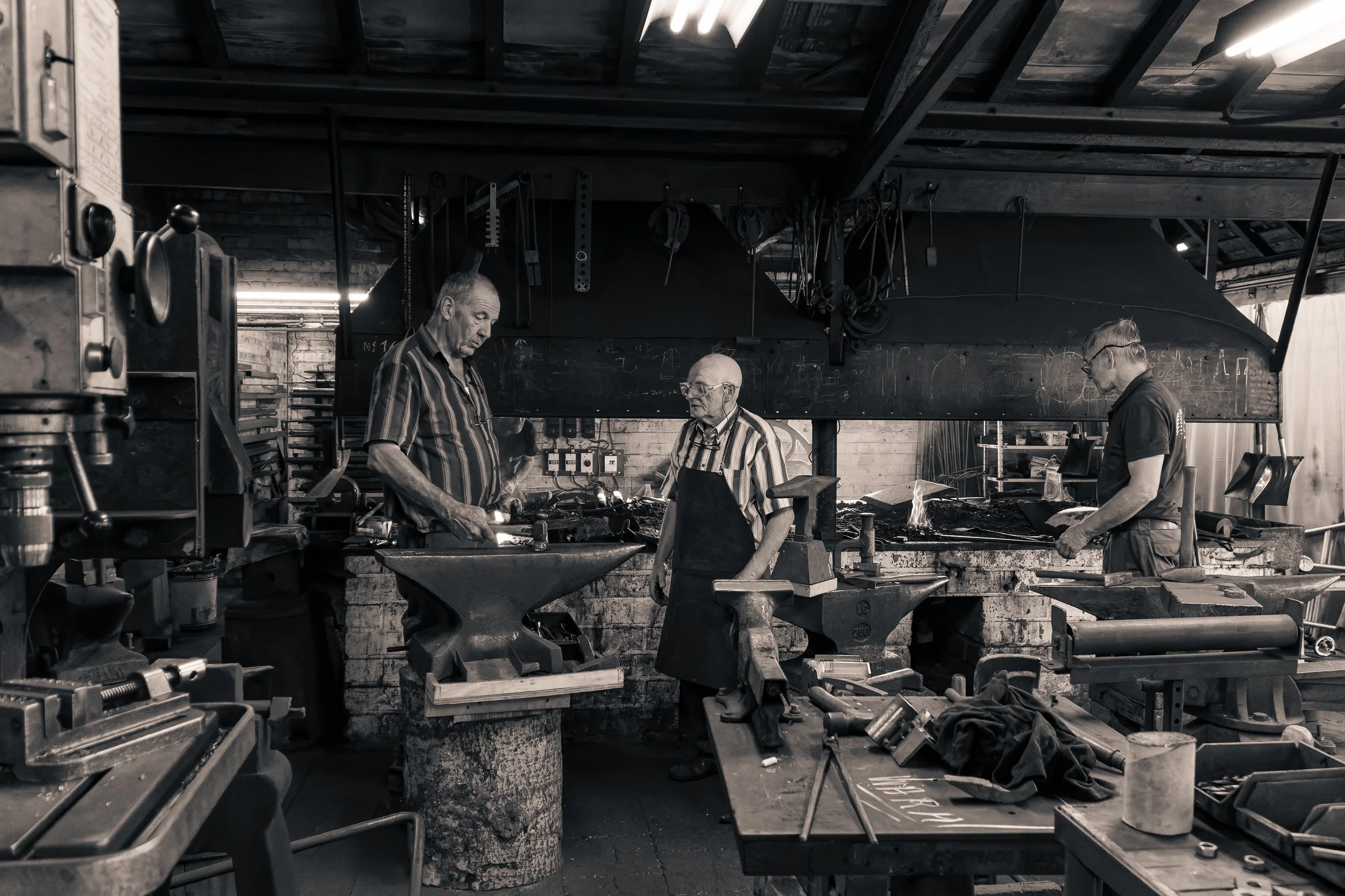 Three men working in a black and white blacksmith workshop with tools and anvils.