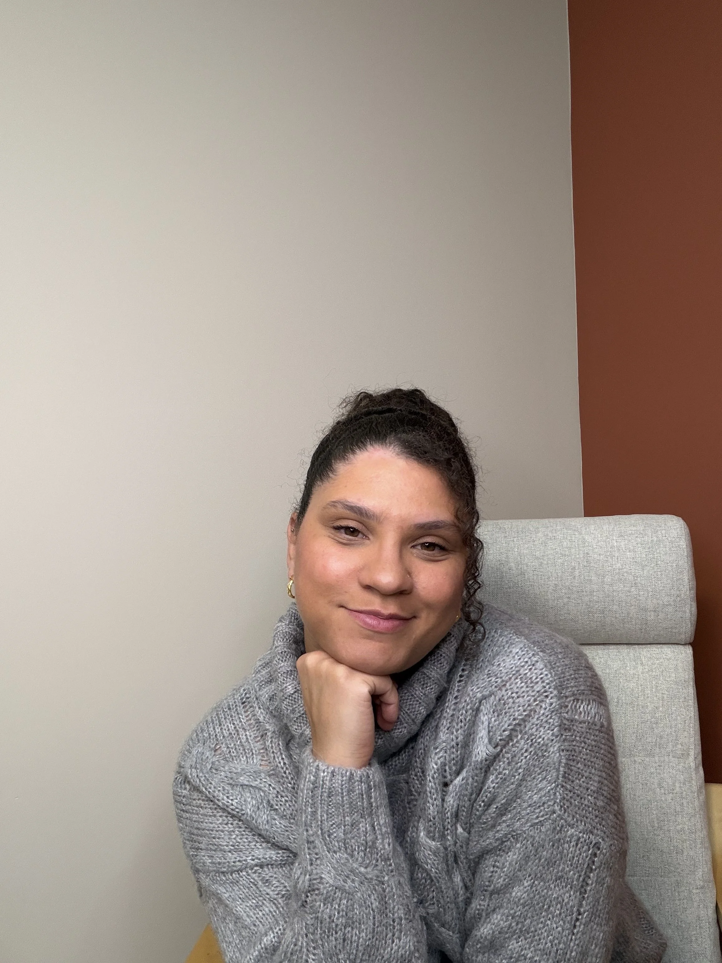 A woman sitting at a desk, wearing a gray sweater, smiling with her chin resting on her hand in an office setting.