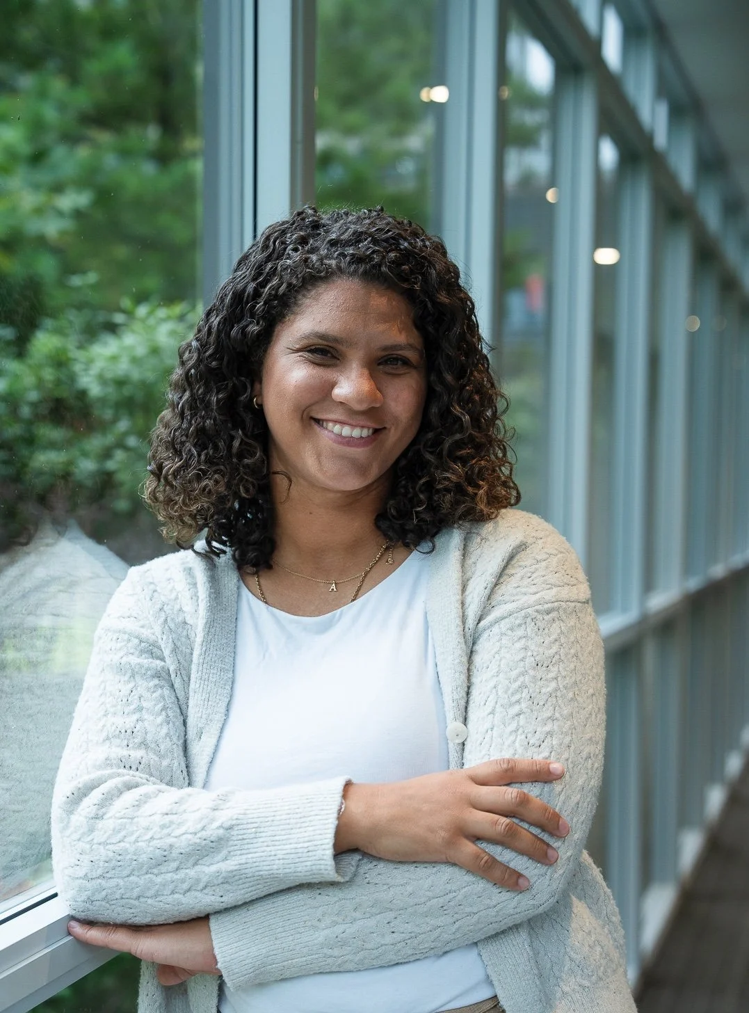 A woman with curly hair smiling, wearing a white shirt and a beige cardigan, standing inside by a large window with green foliage outside.