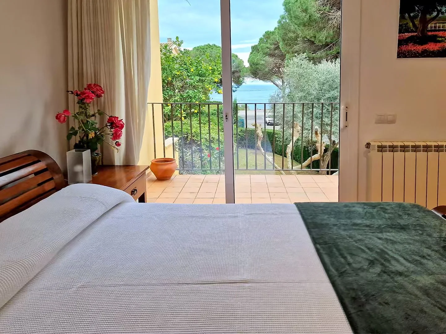 Bedroom with bed, bedside table with pink flowers and a pot, sliding glass door leading to a balcony with view of trees and water.