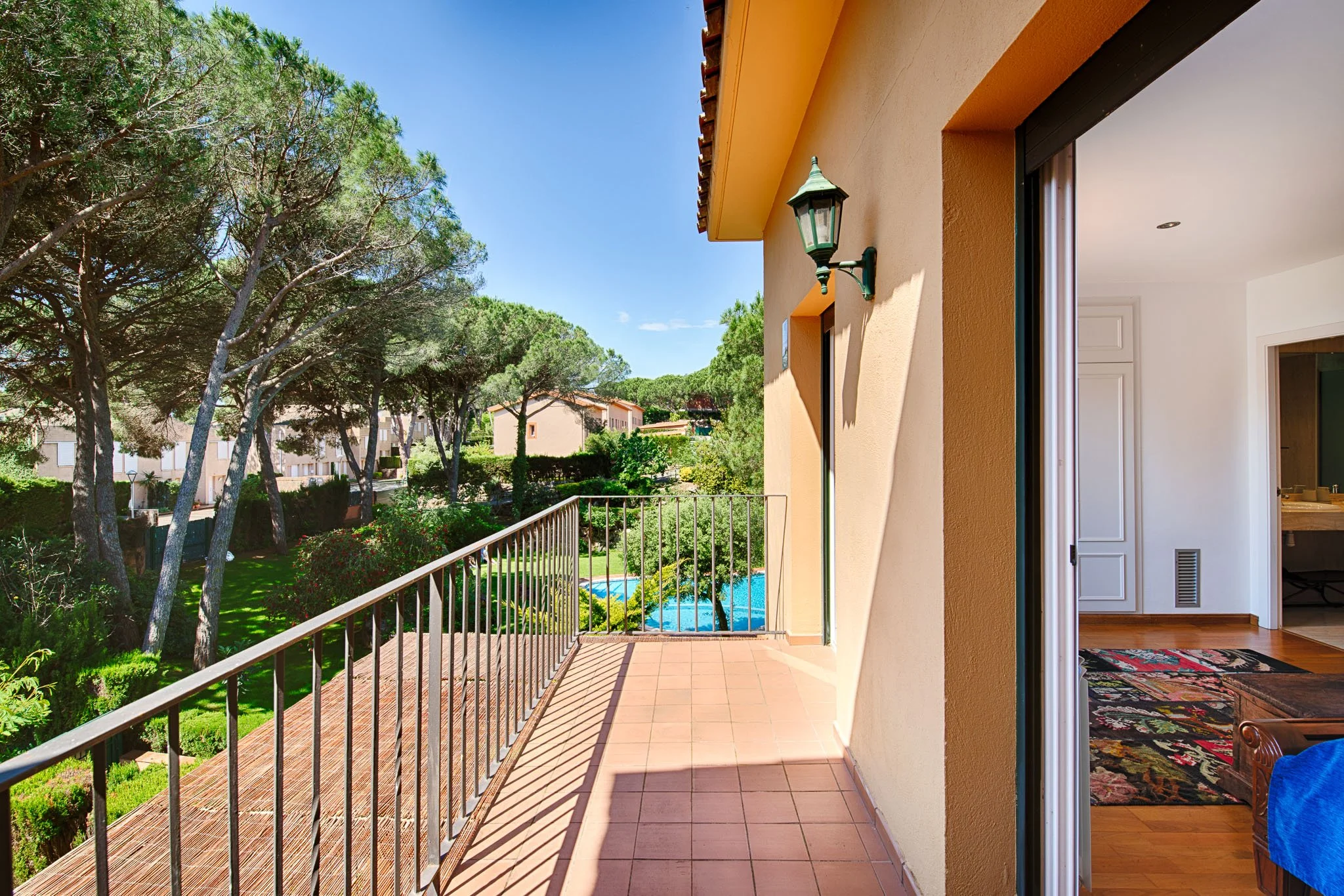 Balcony with a metal railing overlooking a lush green garden with trees and a swimming pool, with the exterior wall of a house and an interior living room visible.