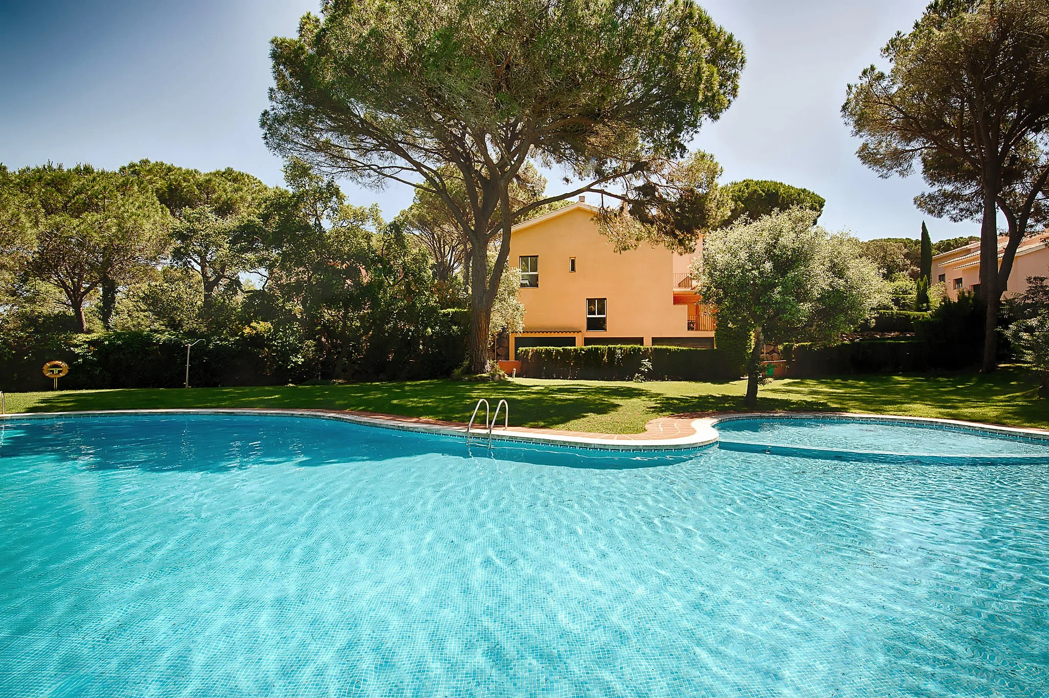 A garden with a swimming pool, surrounded by green grass and tall trees, with a mediterranean house in the background and a clear blue sky.
In Sant Feliu de Guixols In Costa Brava Spain