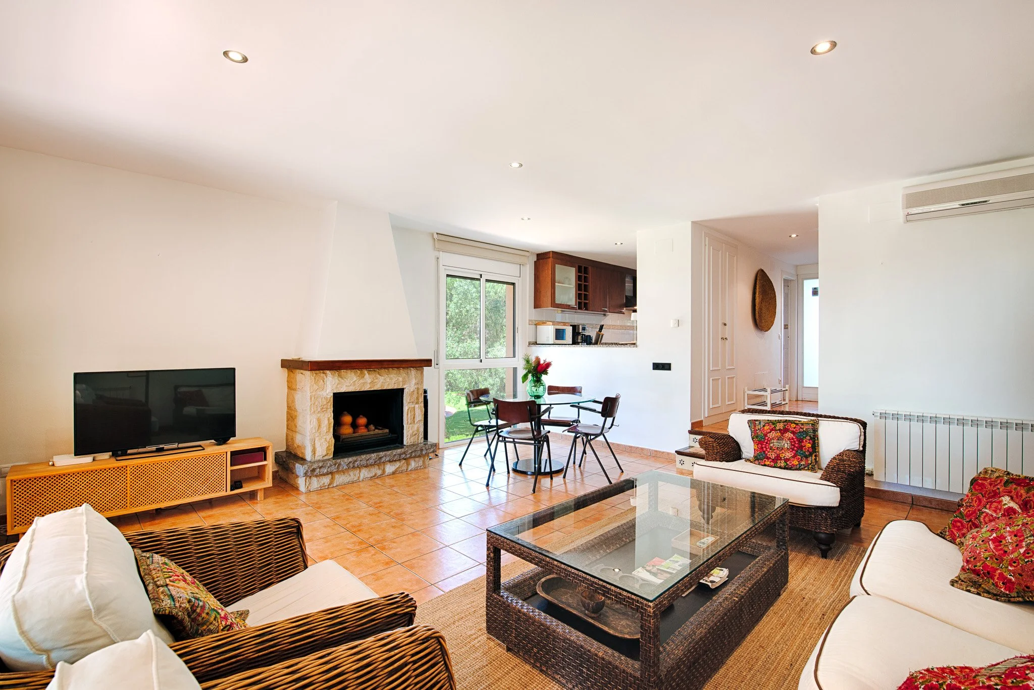 Living room with white walls, tiled floor, and a fireplace. There are couches with floral cushions, a glass coffee table, and a TV on a wooden stand. A dining table with chairs is near the window, and the kitchen with wooden cabinets is visible in the background.