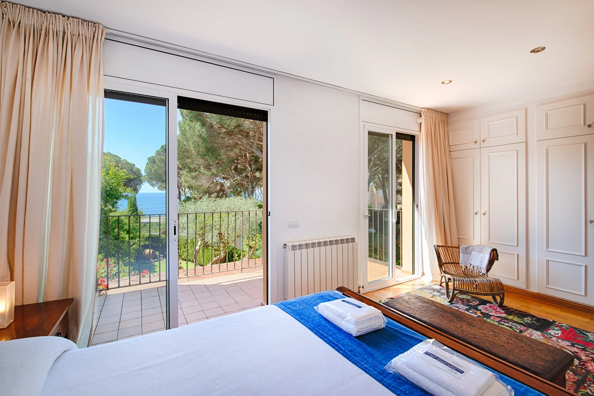 A bedroom with a bed, a wooden desk, a chair, large windows showing greenery and trees, and a balcony with a tile floor.