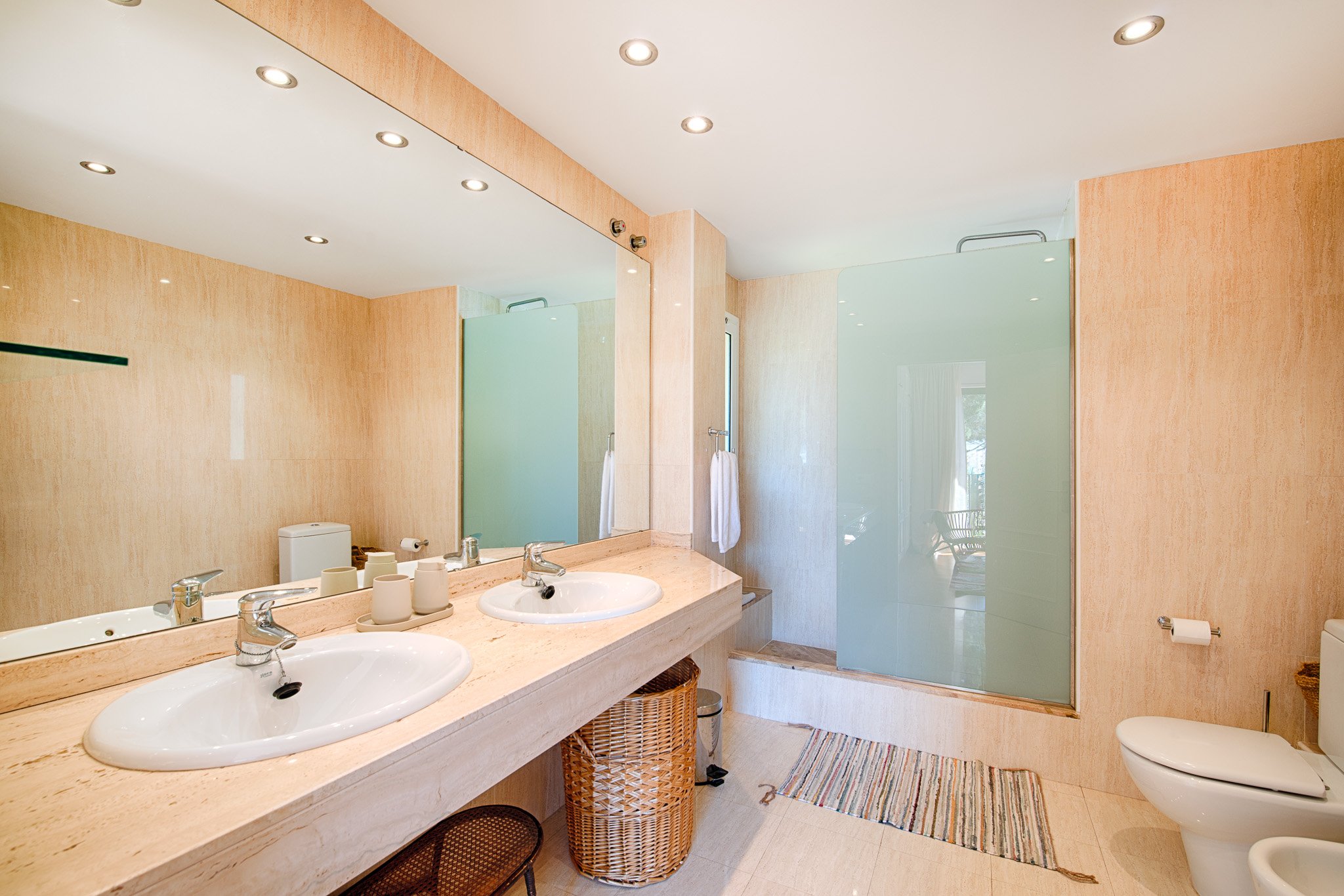 A bathroom with two sinks, a large mirror, beige walls, a glass shower door, a toilet, a striped rug, and various toiletries and storage baskets.