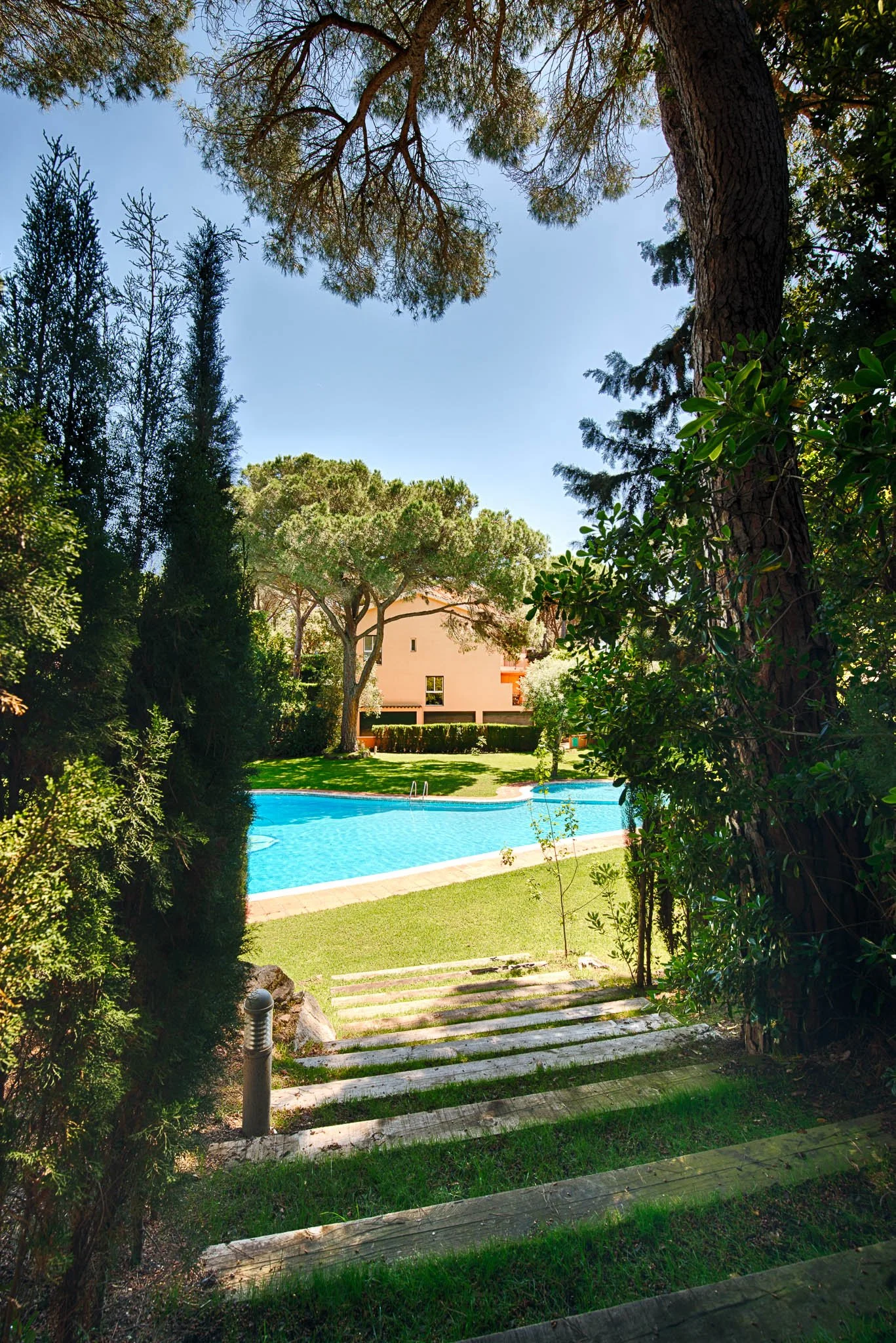 A view of a swimming pool surrounded by a garden in nature. Located in Sant Feliu de Guixols in Costa Brava. The home in the background, and steps leading down to the garden.