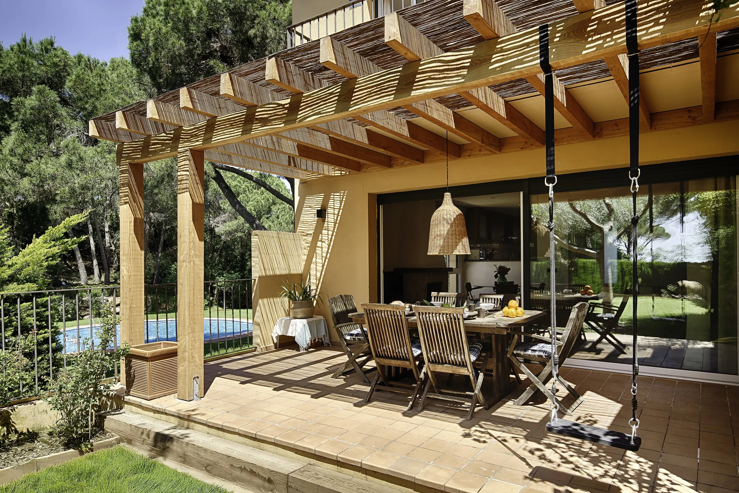 Outdoor Mediterranean terrace with wooden table, chairs, and a swing, overlooking a swimming pool and surrounded by green trees. In Sant Feliu de Guixols In Costa Brava Spain