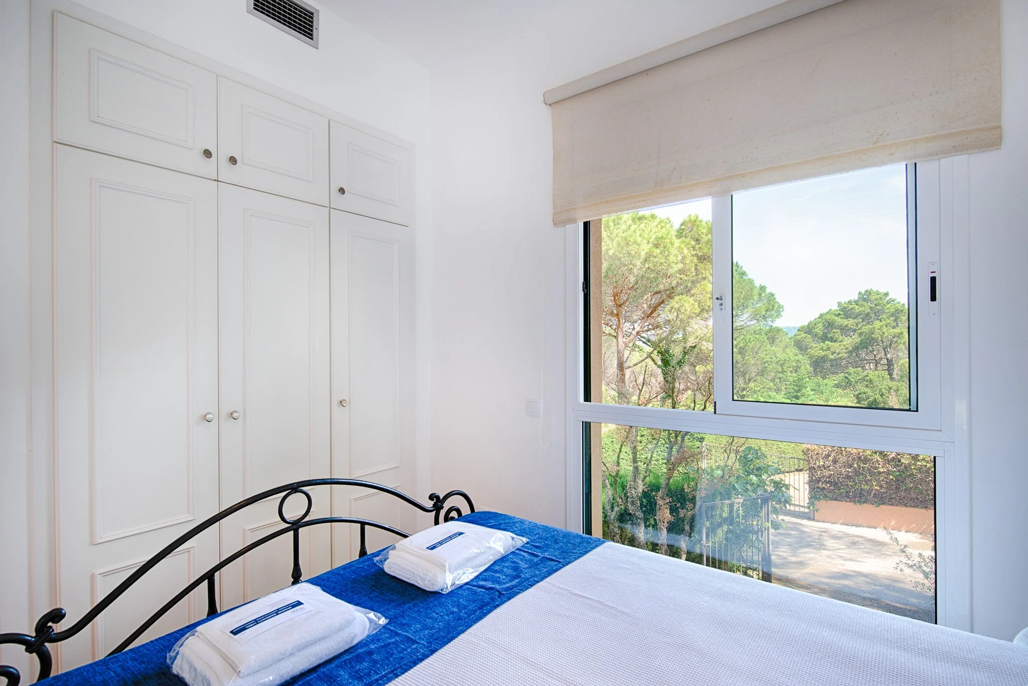 A bedroom with a bed covered in a white sheet and blue bed runner, two pillows wrapped in white packaging, a large window showing trees outside, white built-in cabinets, and a beige window blind.