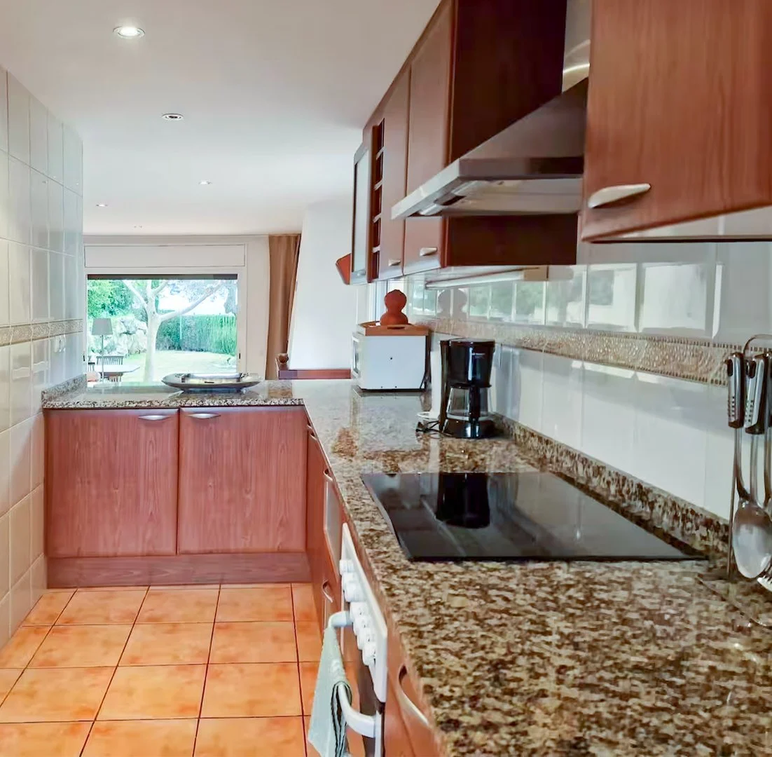 Kitchen with granite countertops, wooden cabinets, a stove, coffee maker, toaster, and a view of a backyard garden with trees through sliding glass doors.