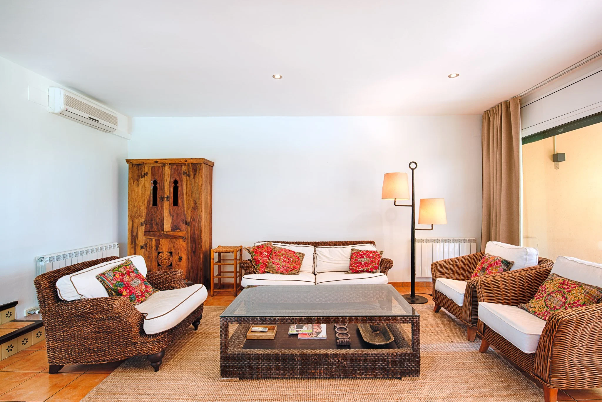 Living room with wicker and wood furniture, colorful cushions, a large window with curtains, and a white wall with air conditioner and lamps.