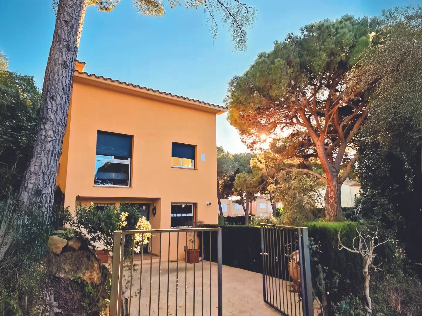 A two-story house painted in a light beige color with two windows on the upper floor and one window and a door on the ground floor. The house is surrounded by tall trees and greenery, with the sun setting behind the trees, casting a warm glow. There is a metal gate at the entrance to the property.
