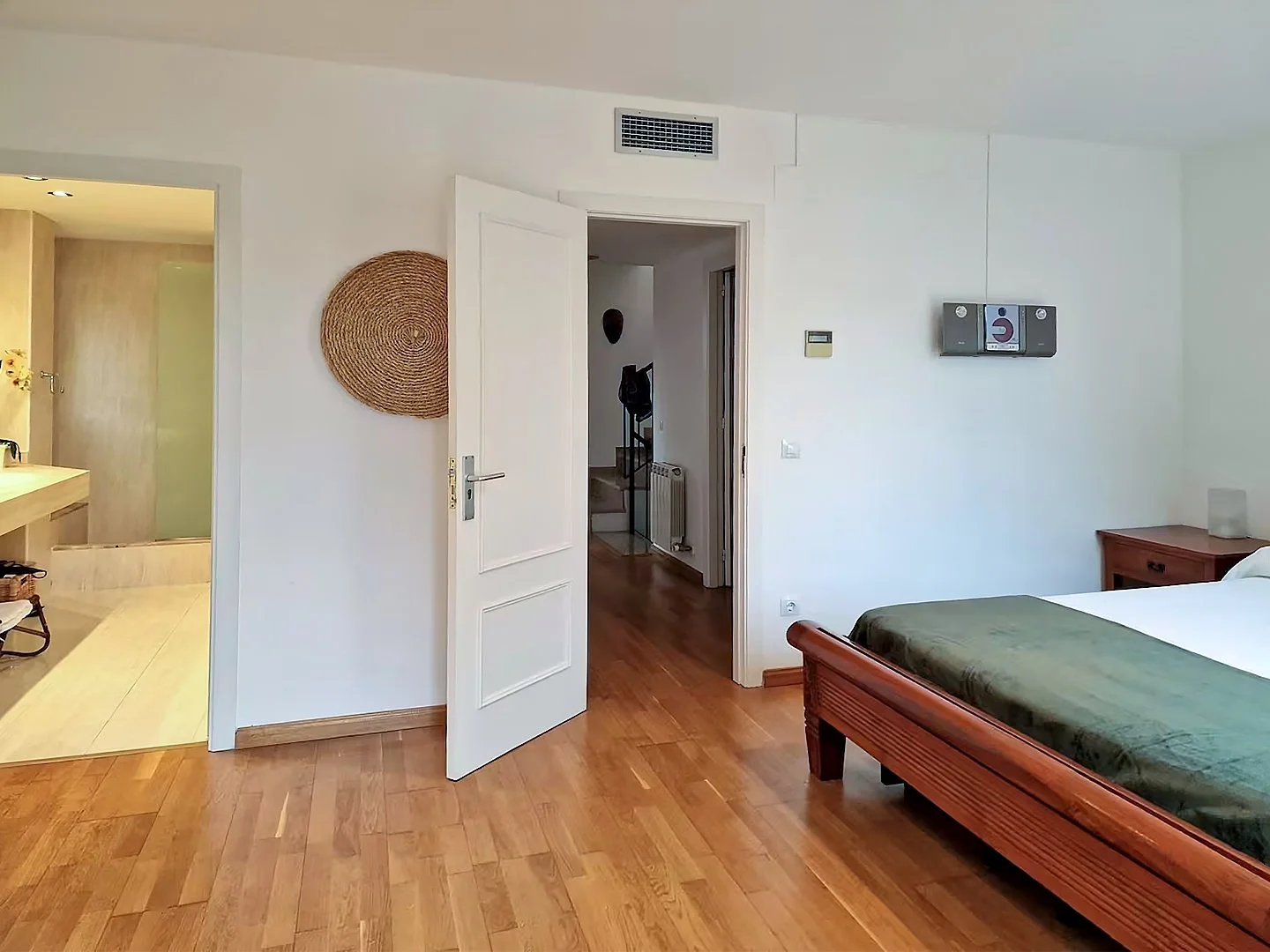 A bedroom with a wooden bed, green bedding, and a side table. An open door reveals a hallway and a staircase. The room has white walls, hardwood flooring, and a wall-mounted TV.