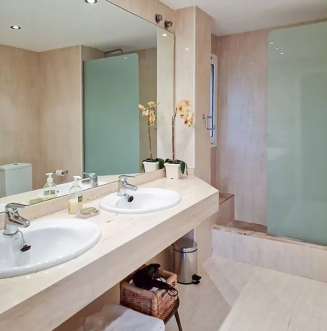 Bathroom with dual sinks, large mirror, orchid plants, toiletries, shower area with frosted glass door, and beige tiled walls and floor.