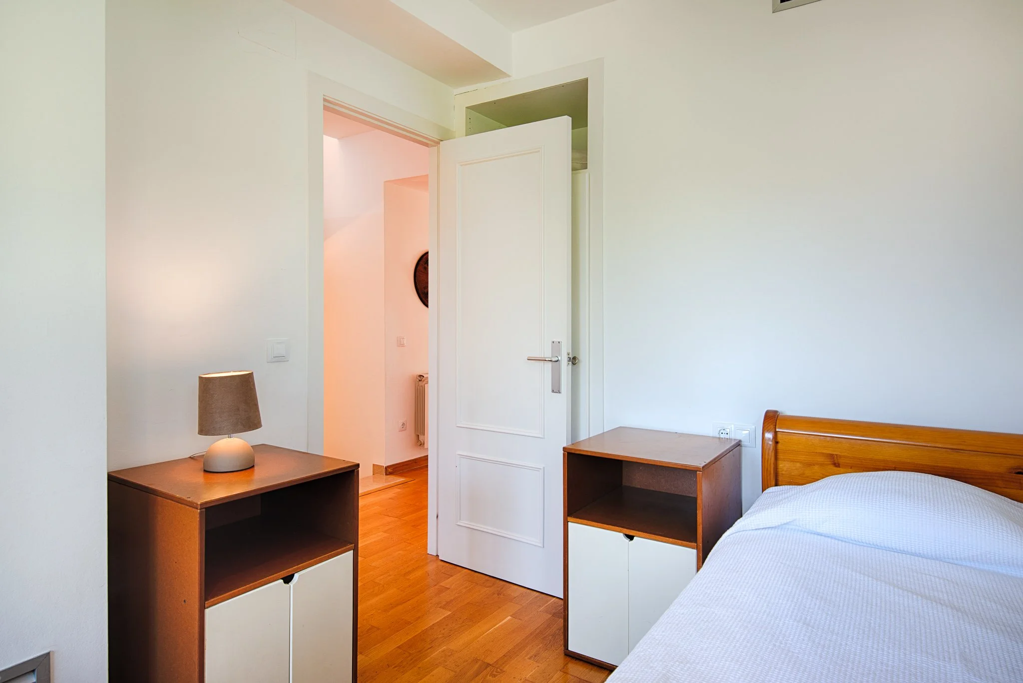 Interior of a bedroom with a bed, two wooden nightstands, a lamp, and an open door leading to a hallway.
