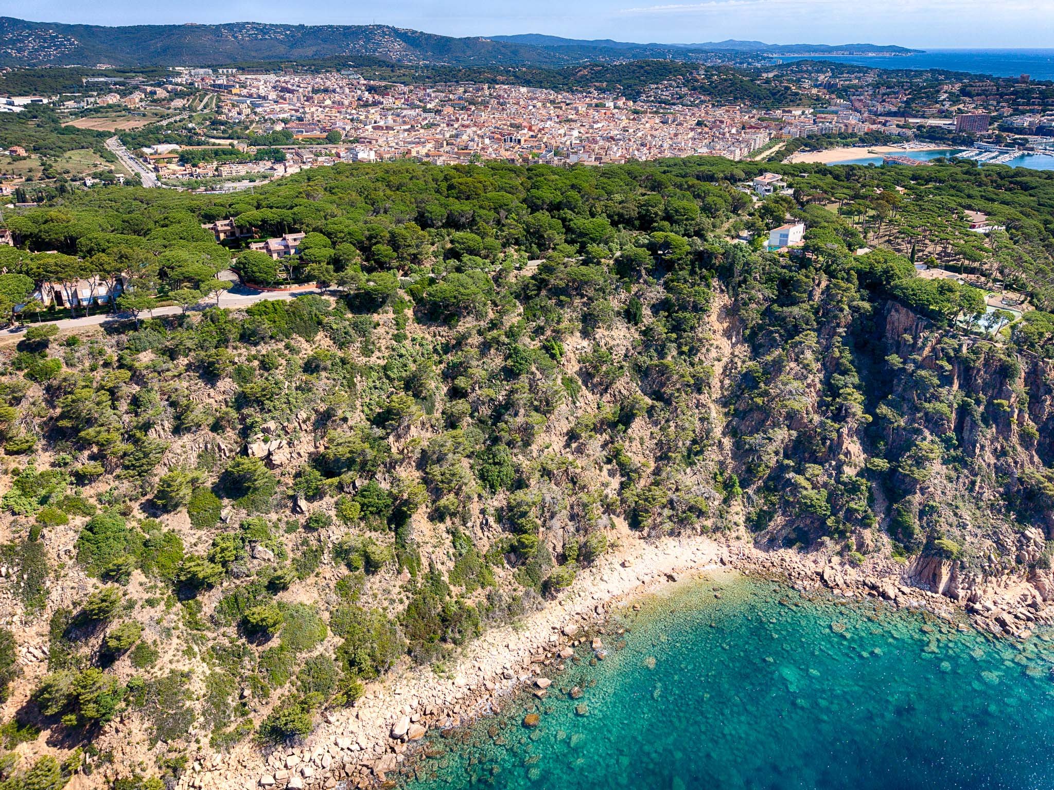 Aerial view of houses in Sant Feliu de Guixols, Les Penyes surrounded by forest near the sea line, showing the village extending into the distance with mountains in the background. In Costa Brava, Spain