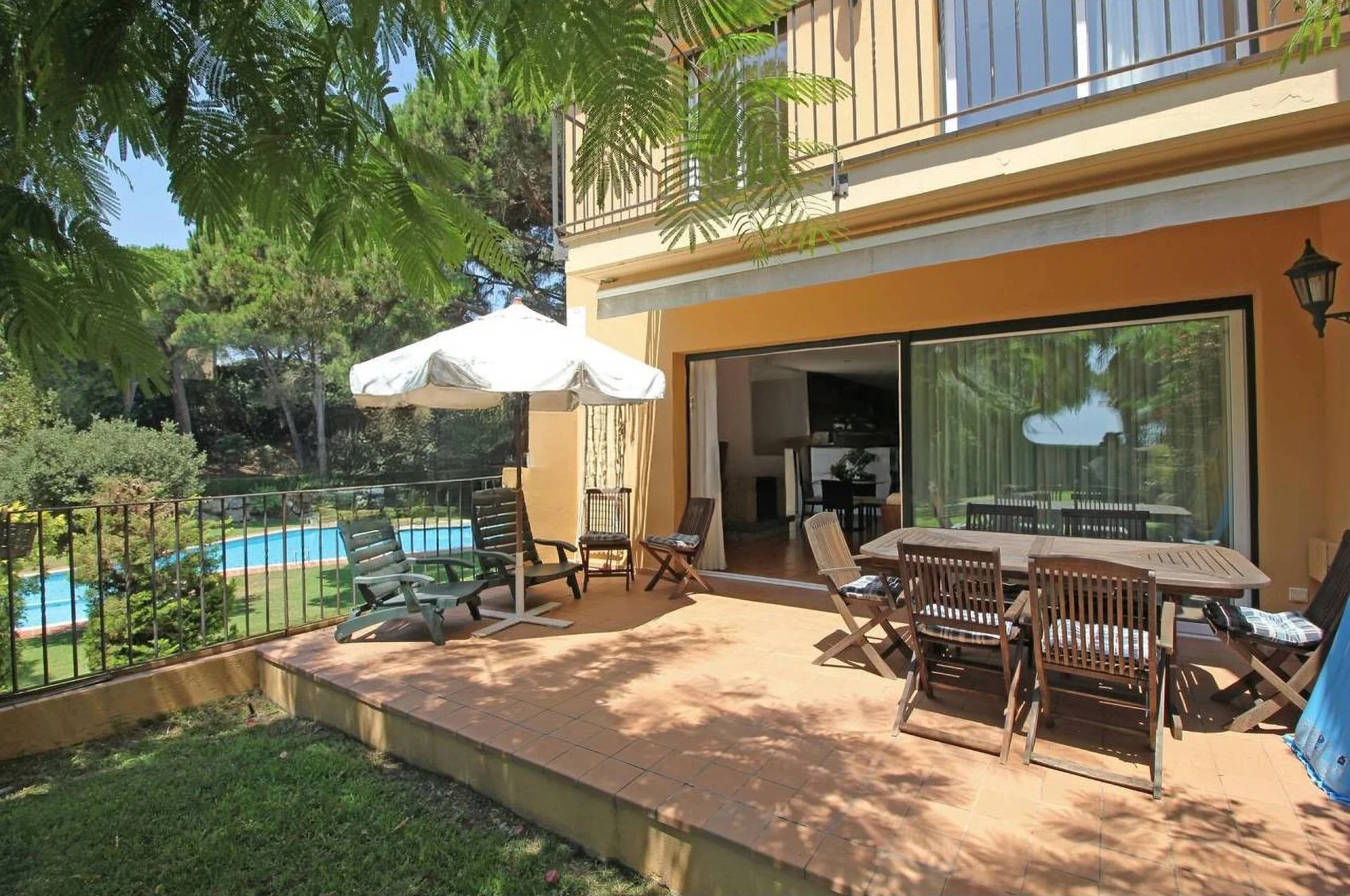A patio with outdoor furniture, including a table and chairs, a white umbrella, and a pool in the background surrounded by greenery.