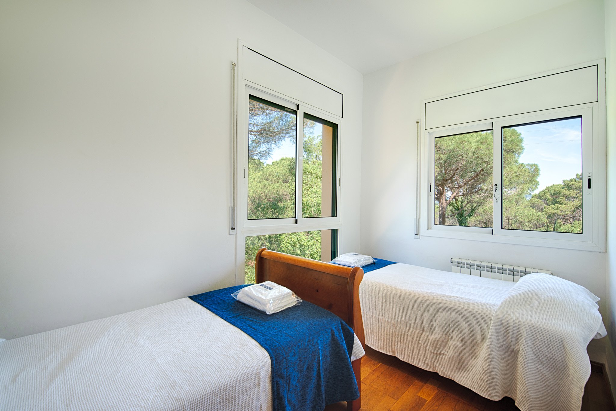 A bedroom with two single beds, each covered with white sheets, blue bed runners, and pillows. The room has large windows showing green trees outside and natural light coming in.