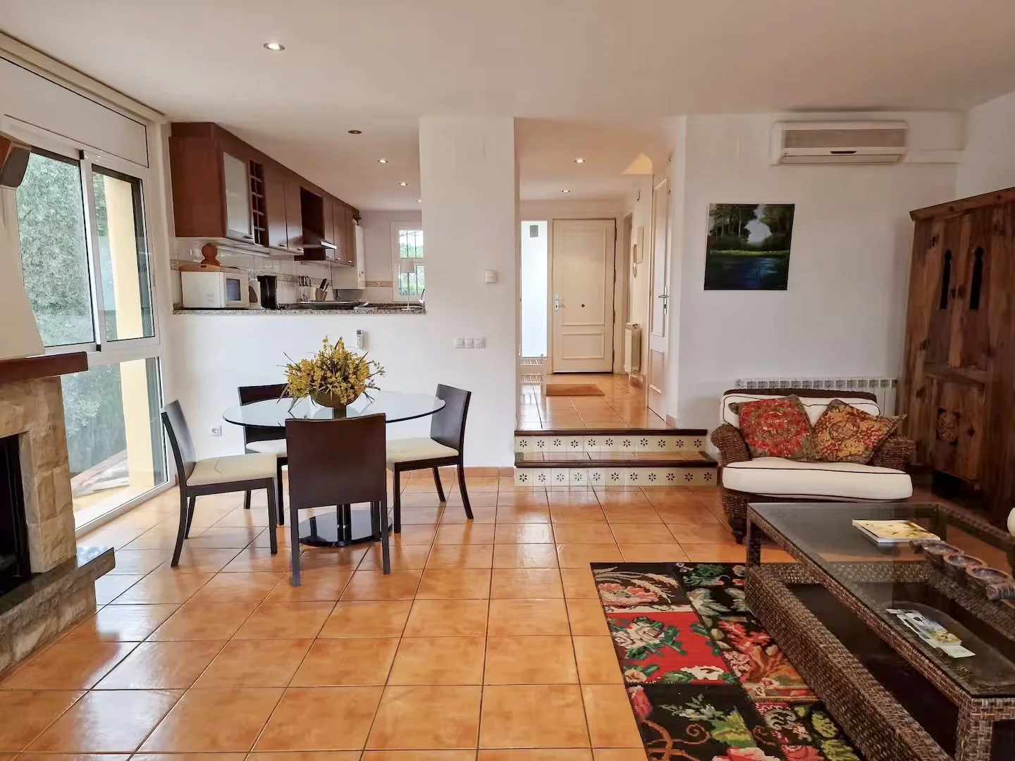 Living room with tiled floor, white walls, wicker furniture with colorful cushions, floral rug, dining table with four chairs and a floral centerpiece, open kitchen with wooden cabinets and countertop, staircase, and artwork on the wall.