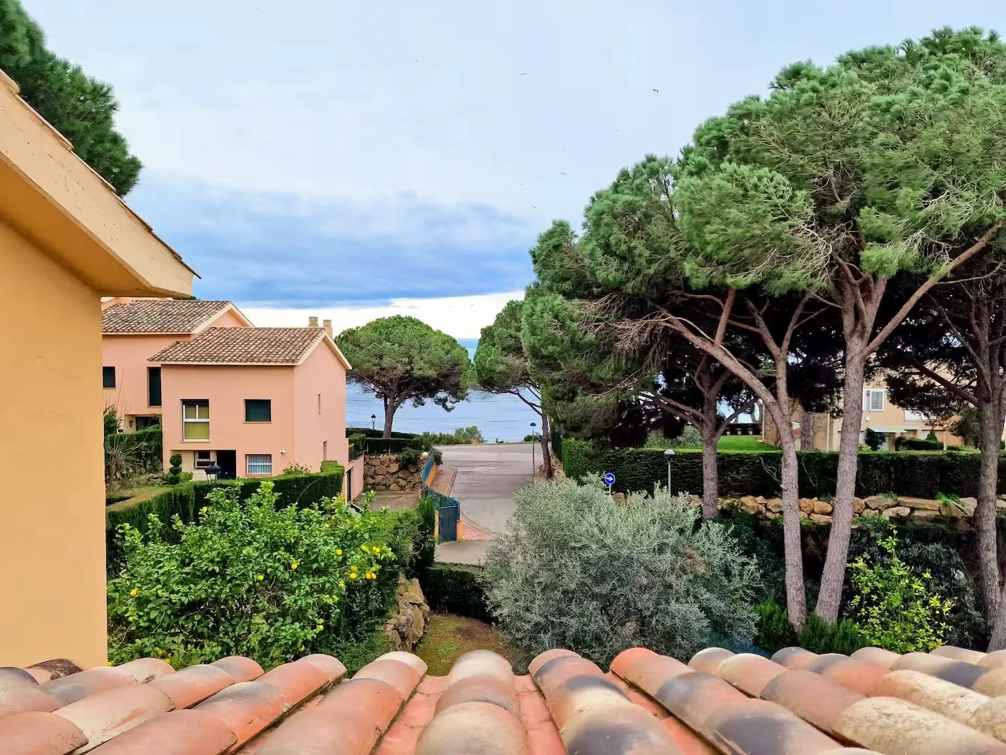 View from a balcony with terracotta roof tiles looking over a residential area with pastel-colored buildings, green trees, bushes, and a glimpse of the ocean in the distance under a partly cloudy sky.