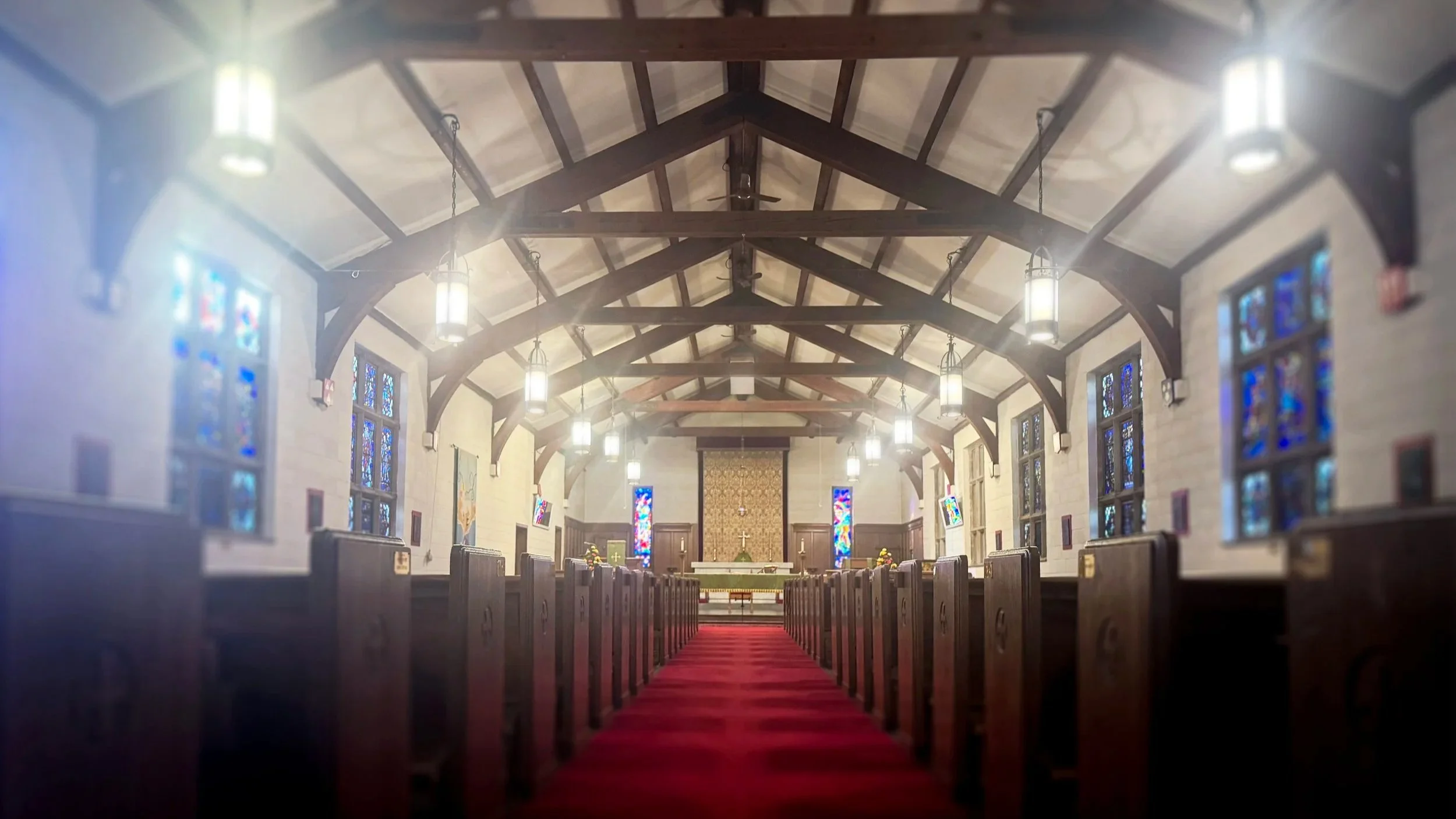 Interior view of a church sanctuary with wooden pews, stained glass windows, and hanging lights, leading up to the altar at the front.