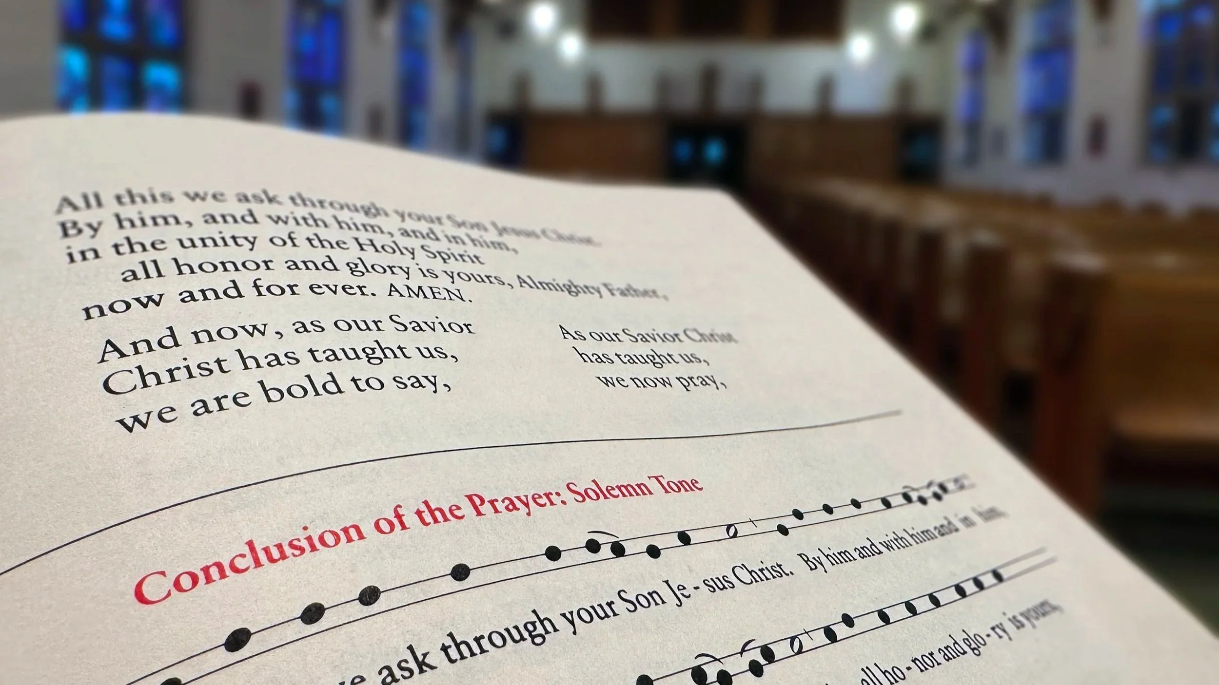 Close-up of a printed page with religious text, set inside a church with tall stained glass windows and wooden pews.