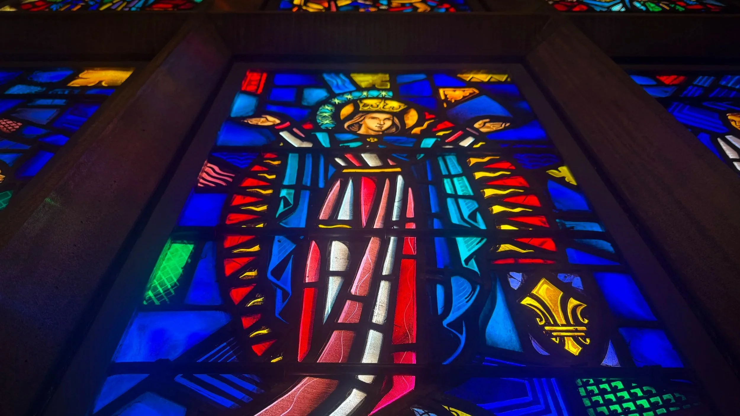 Colorful stained glass windows depicting a haloed figure with raised hands, surrounded by various geometric patterns and vibrant colors.