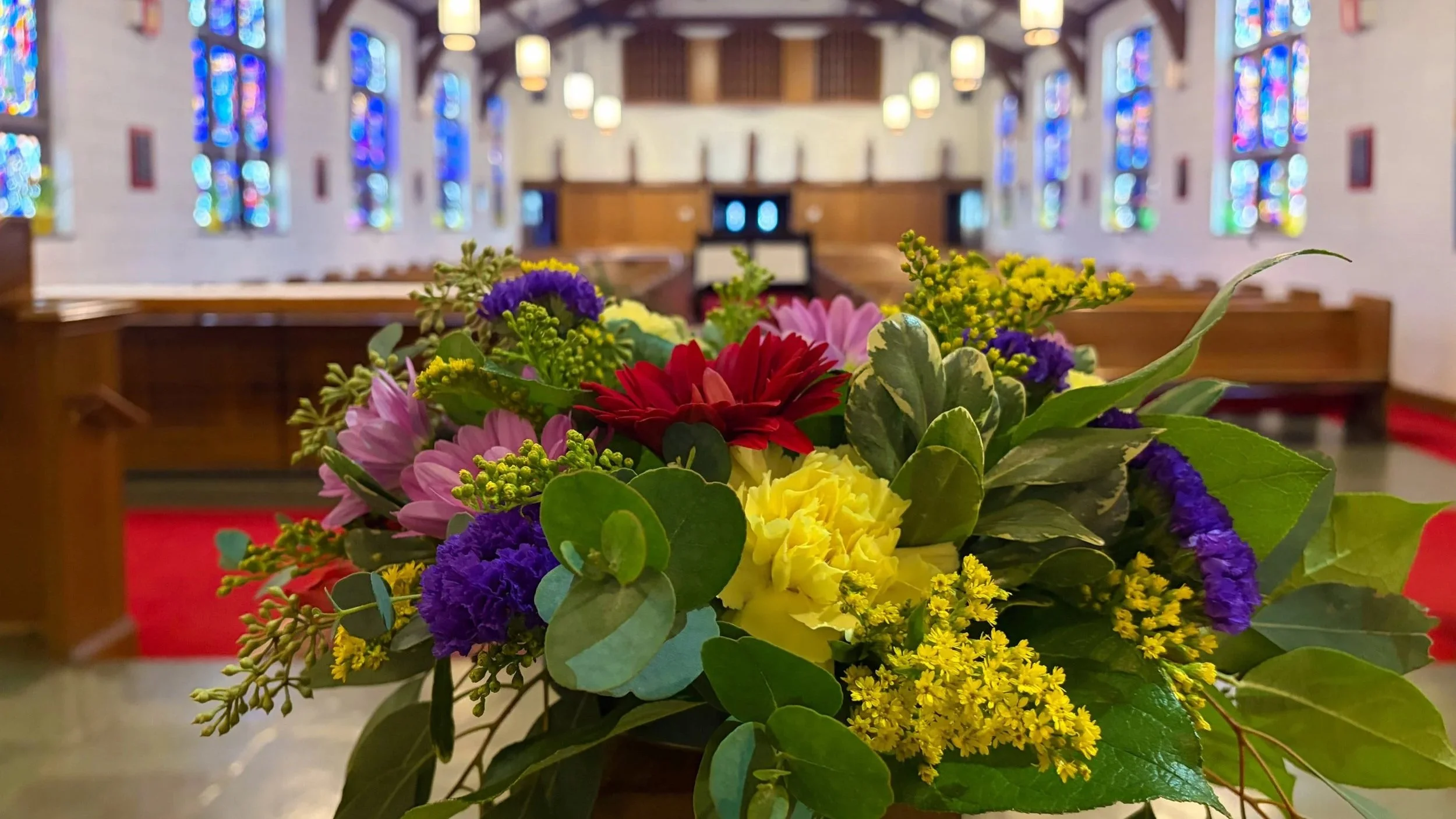 A colorful bouquet of flowers placed on a wooden pew inside a church with stained glass windows and hanging lights.