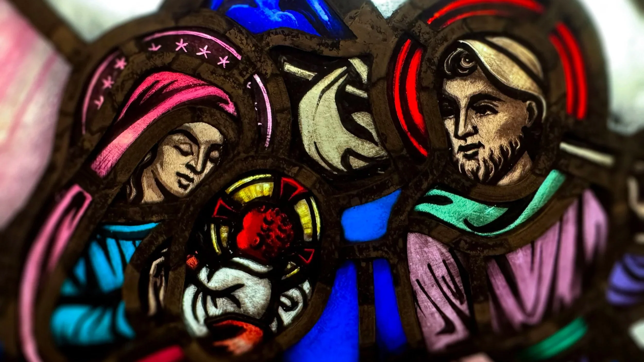 Close-up of a colorful stained glass art featuring three human faces with diverse features, colorful patterns, and vibrant hues including red, blue, pink, green, and yellow.