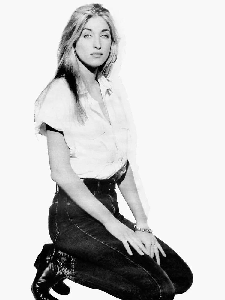 Black and white photo of a young woman with long hair, wearing a short-sleeved shirt, high-waisted jeans, and boots, sitting on her knees against a plain background.