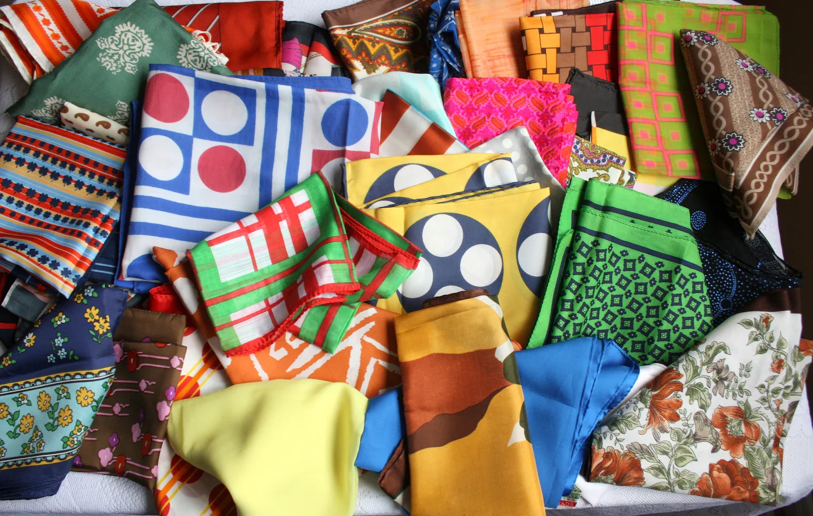 A collection of colorful bandanas with various patterns including floral, geometric, and abstract designs, arranged haphazardly on a surface.