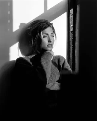 A woman with short hair sitting by a window, gazing out in sunlight, wearing a sweater, in black and white.