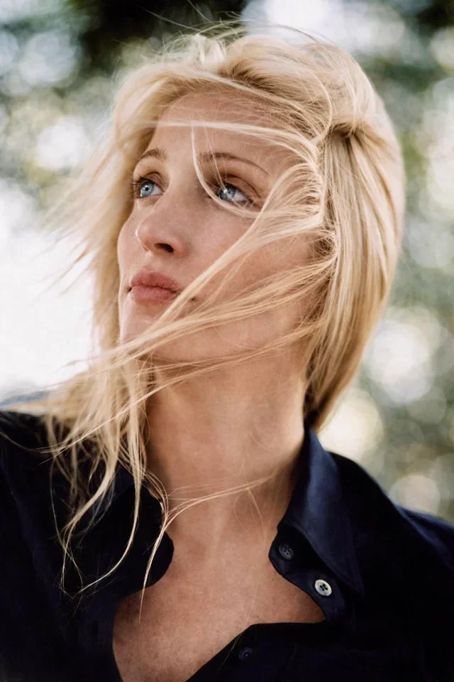 A woman with blonde hair looking off to the side, outdoors, with wind-blown hair and a blurred background of trees.