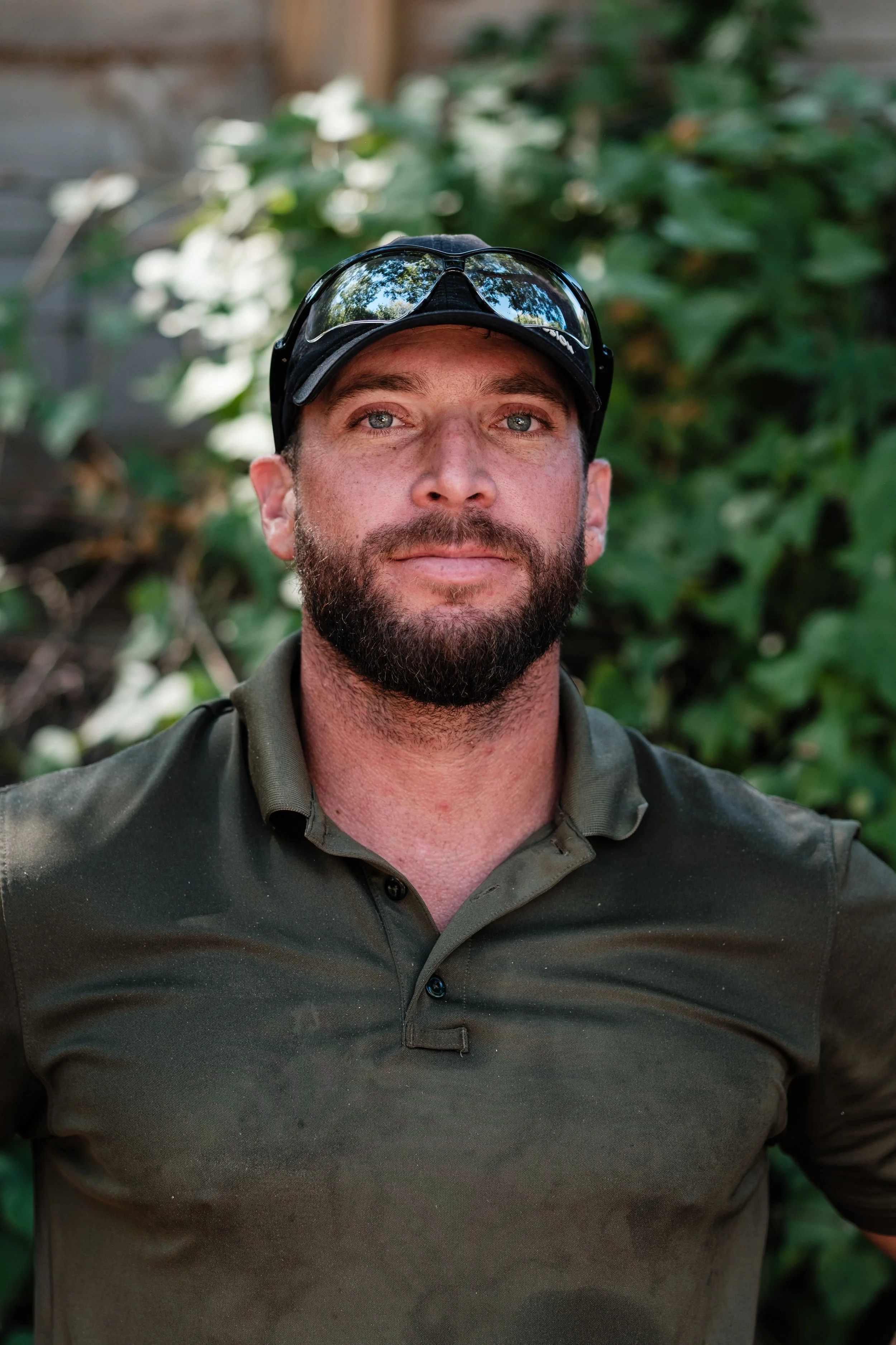 A man with a beard and mustache wearing a dark green polo shirt and sunglasses, standing outdoors with greenery in the background.