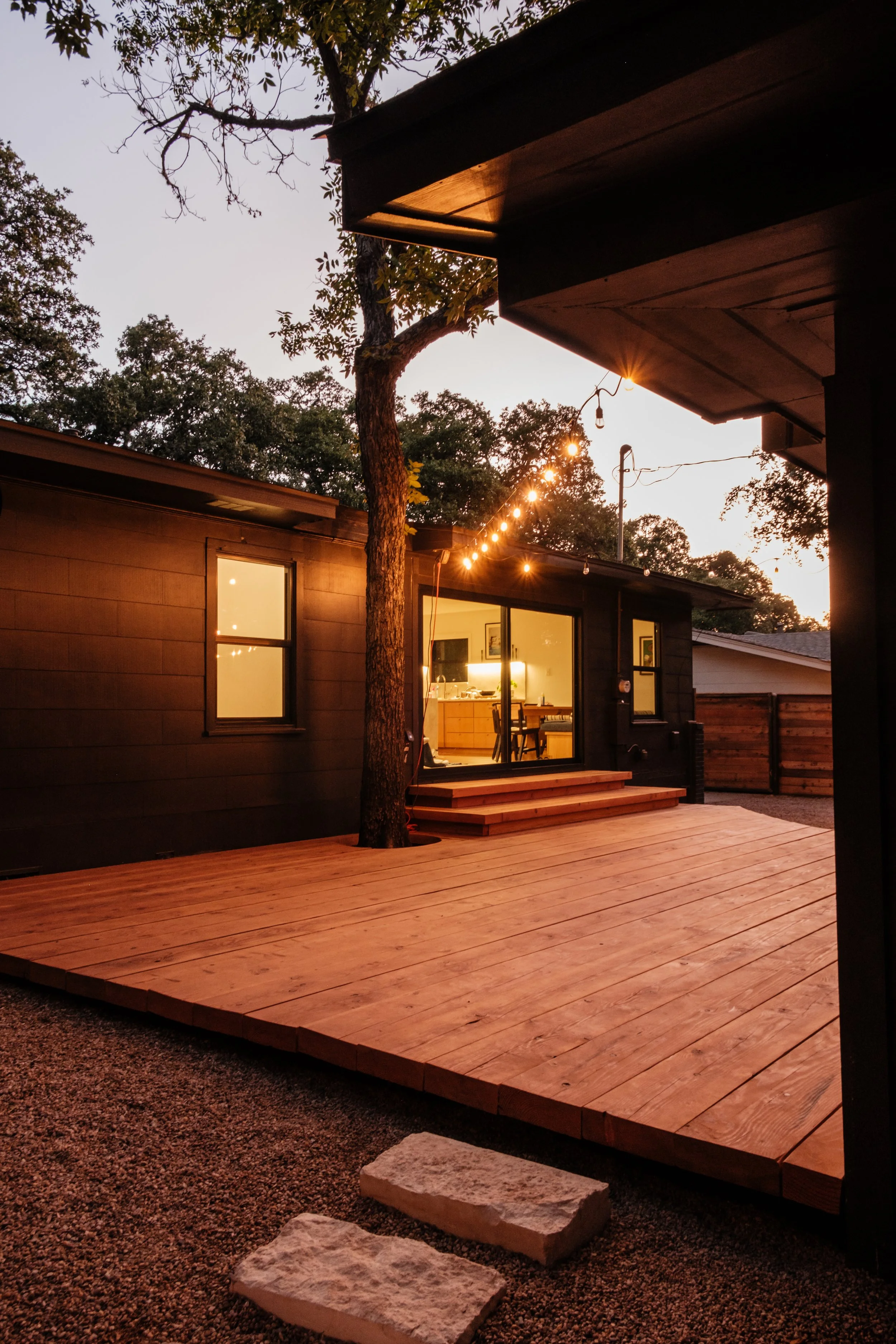 A backyard with a wooden deck, lit string lights, and an exterior view of a house through sliding glass doors showing a warmly lit interior kitchen and dining area. There's a tree positioned near the house and stone steps leading onto the deck, all during twilight.