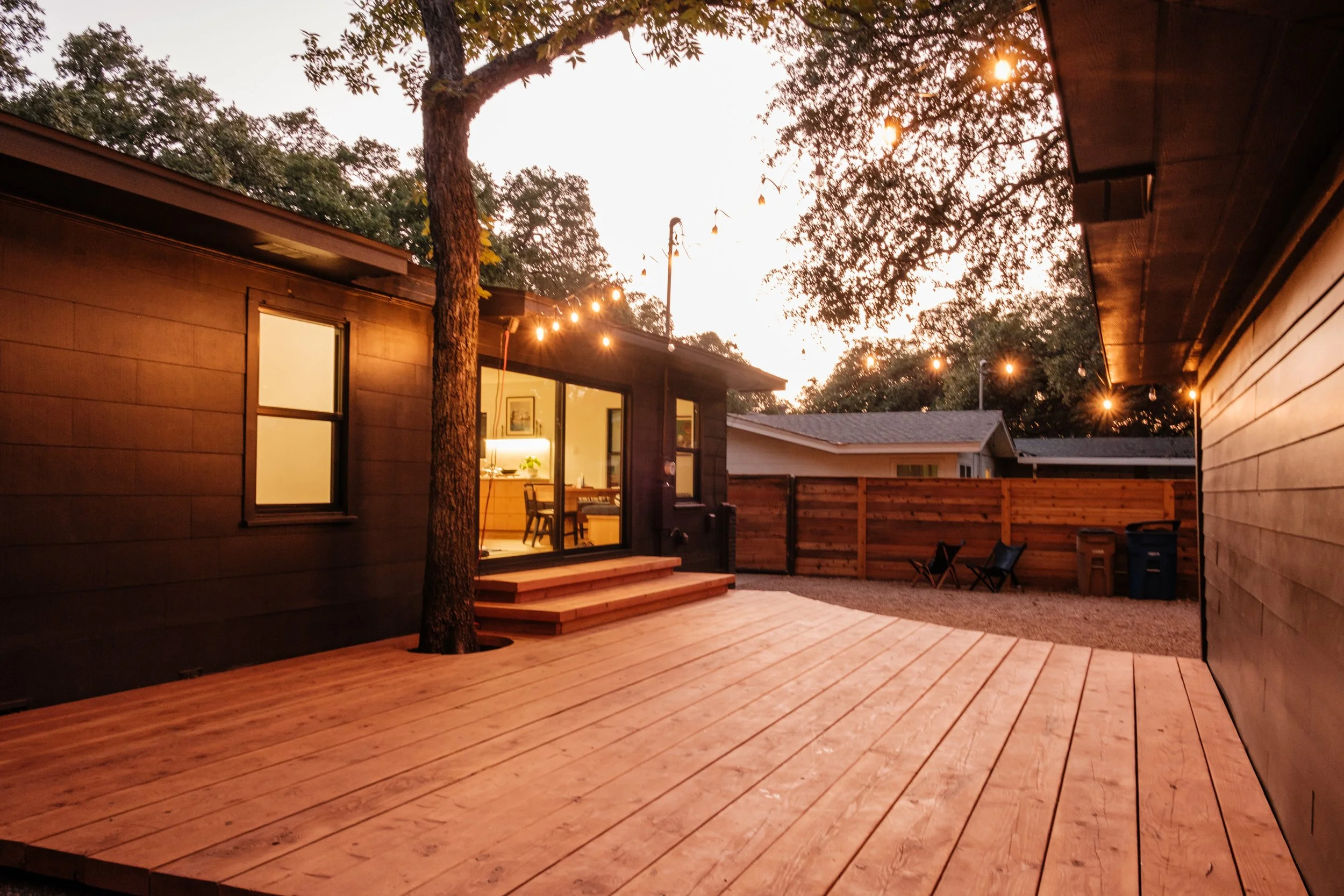 A backyard deck with wooden flooring, outdoor string lights, trees, and a neighboring house in the background at sunset.