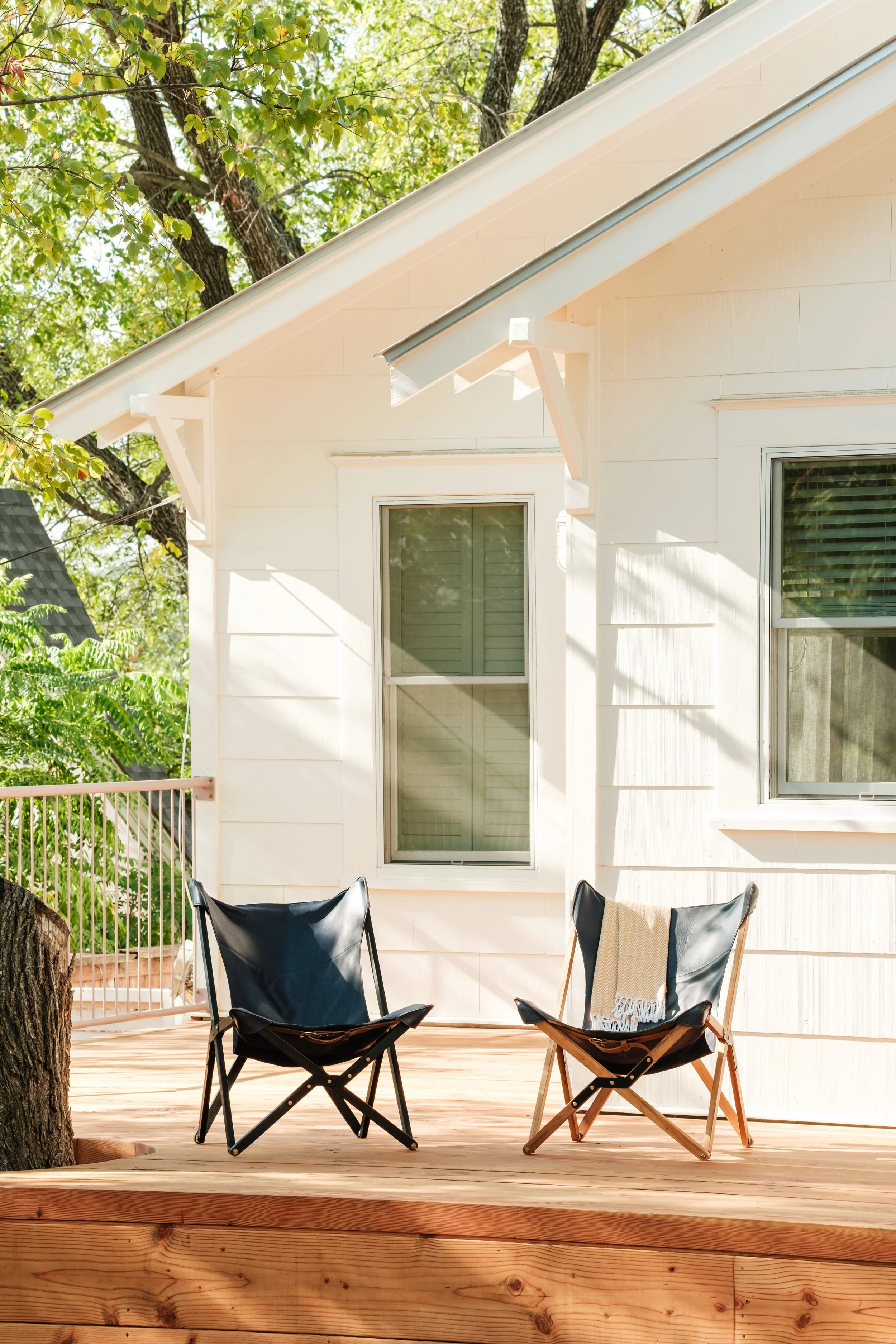A wooden deck with two black and brown folding chairs, one with a beige throw blanket, in front of a white house with large windows and green trees in the background.