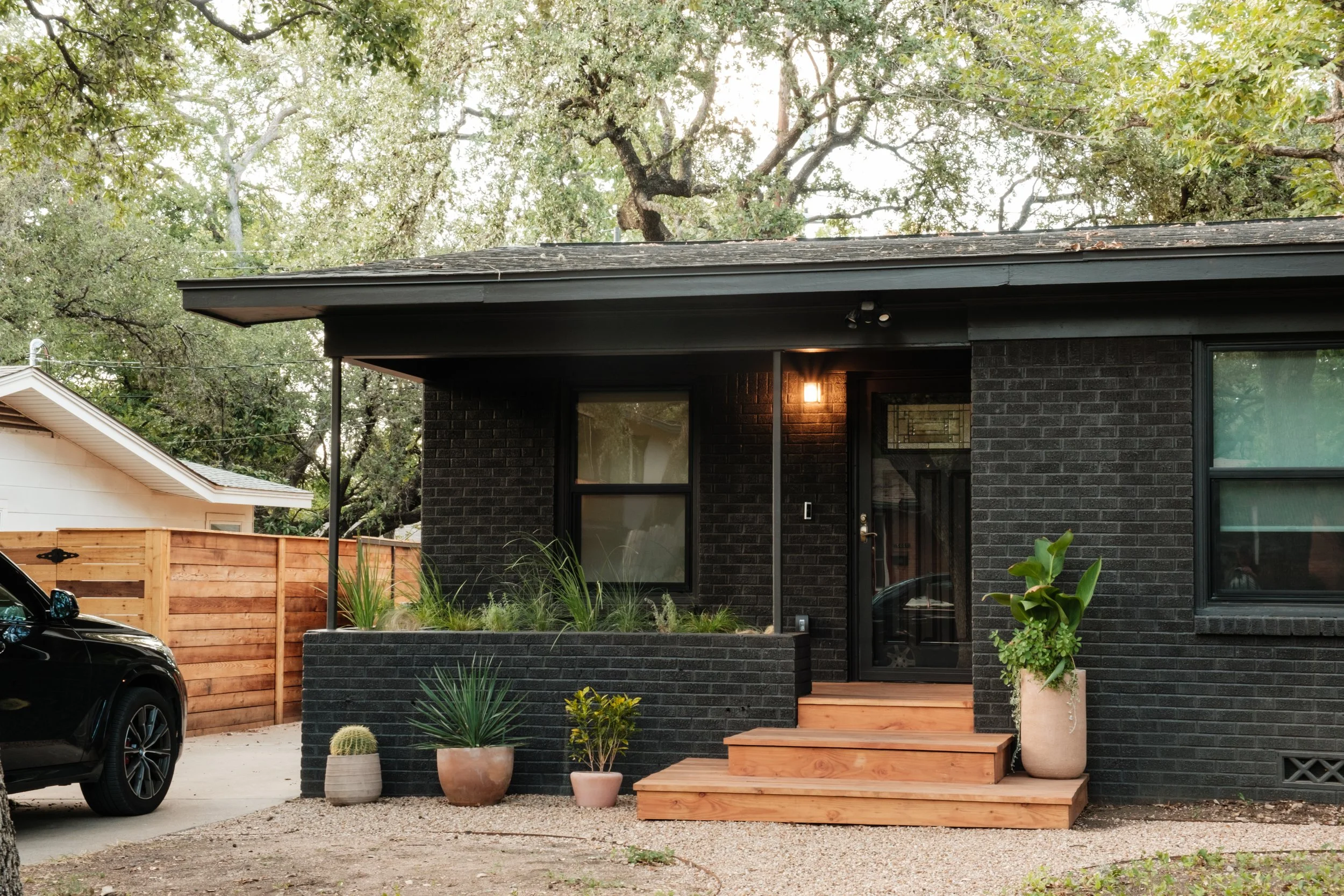 A modern house with black brick exterior, wooden steps leading to the front door, and potted plants including a large leafy plant in a white pot. There is a black car parked on the driveway and a wooden fence on the side, with trees in the background.