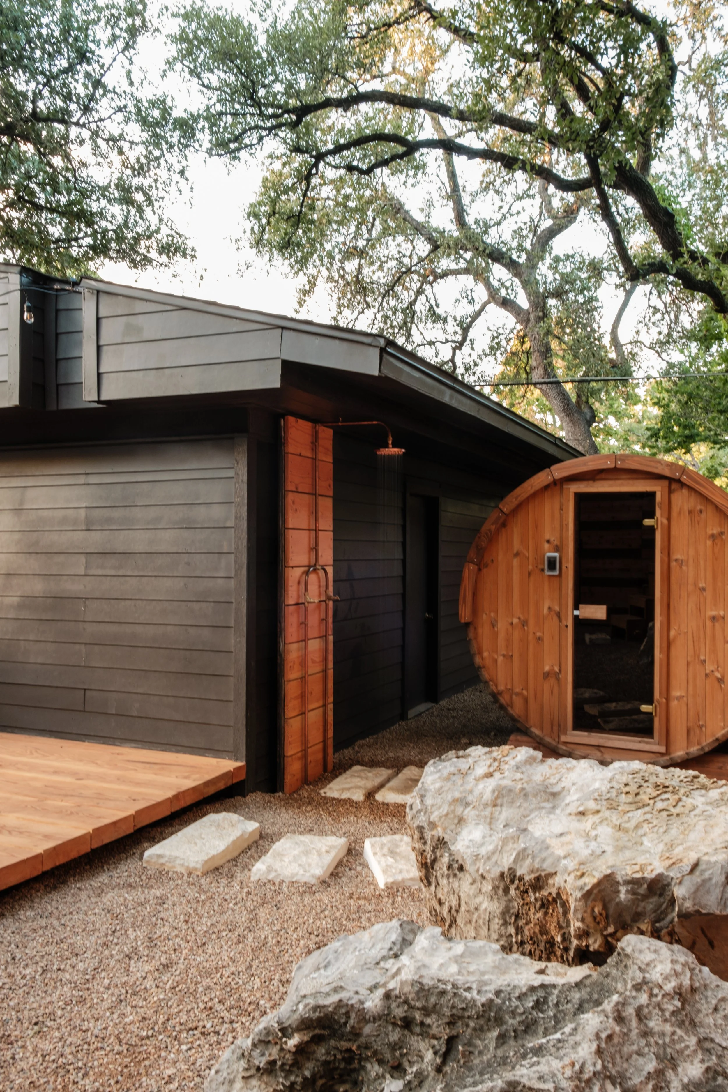 Outdoor wooden sauna with a glass door and a small outdoor shower, surrounded by rocks and gravel, with trees in the background.