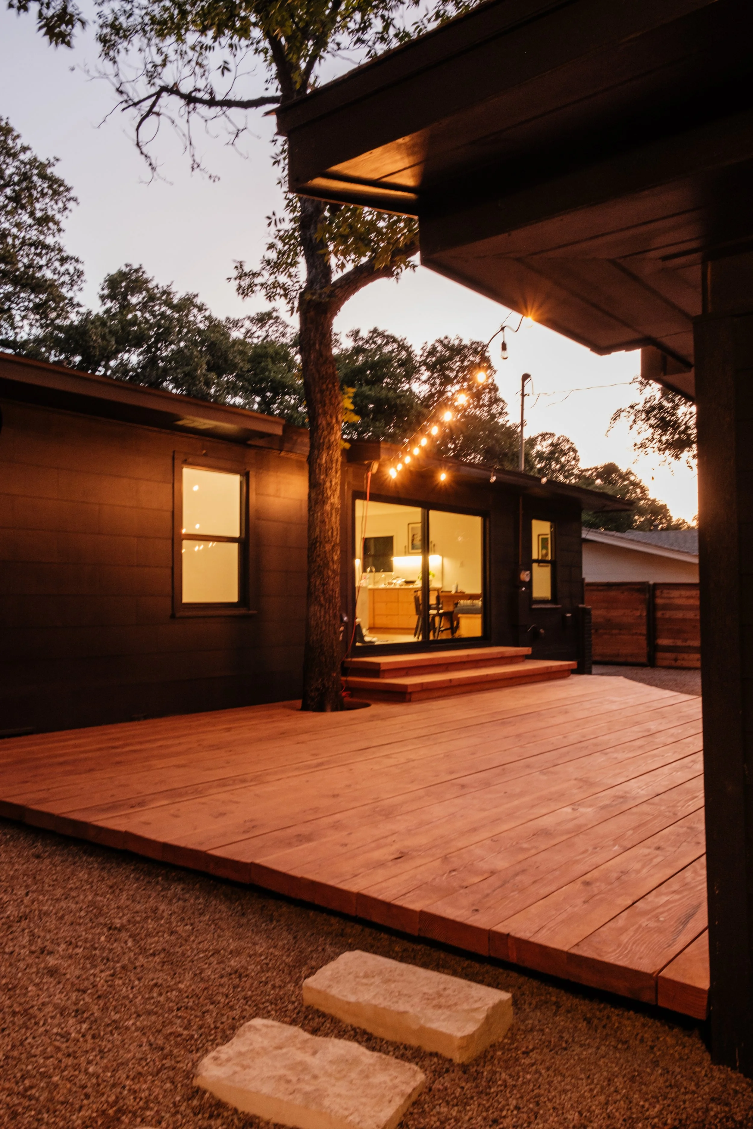 View of a backyard with a wooden deck, string lights, a tree growing through the deck, and a house with illuminated interior visible through sliding glass doors at sunset.