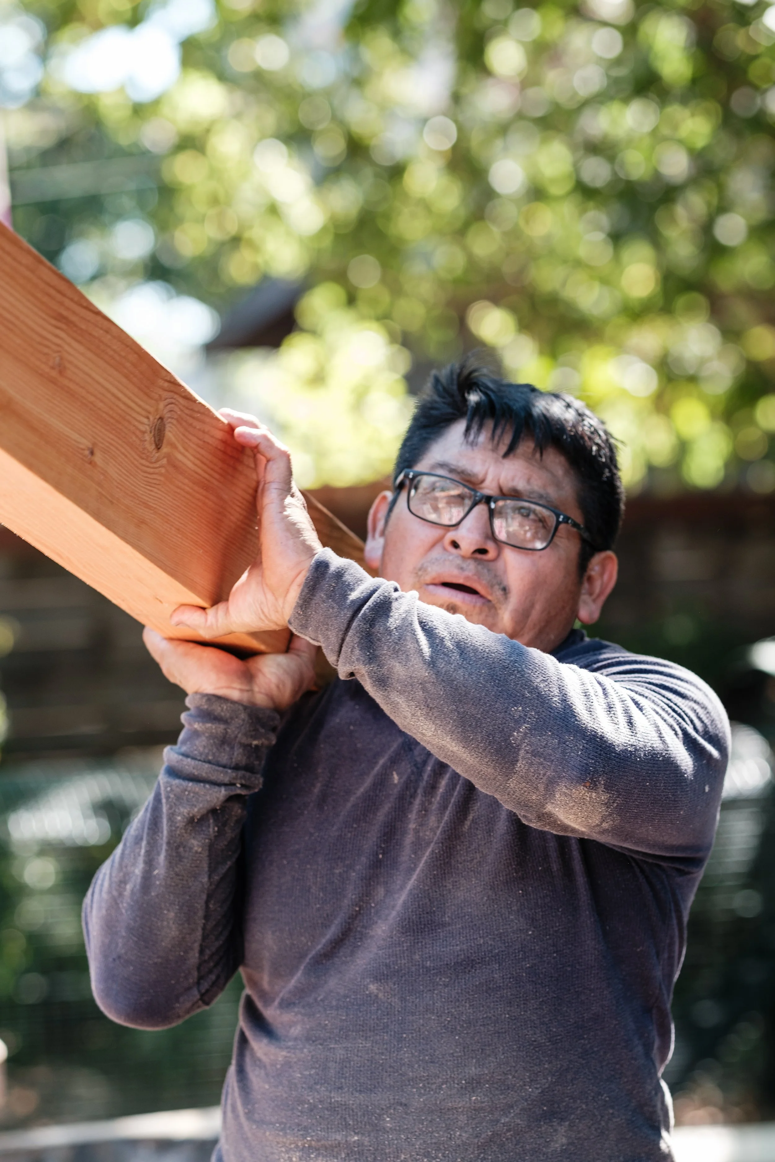 A man wearing glasses and a dirty long-sleeve shirt looks worried as he holds a wooden plank on his shoulder.