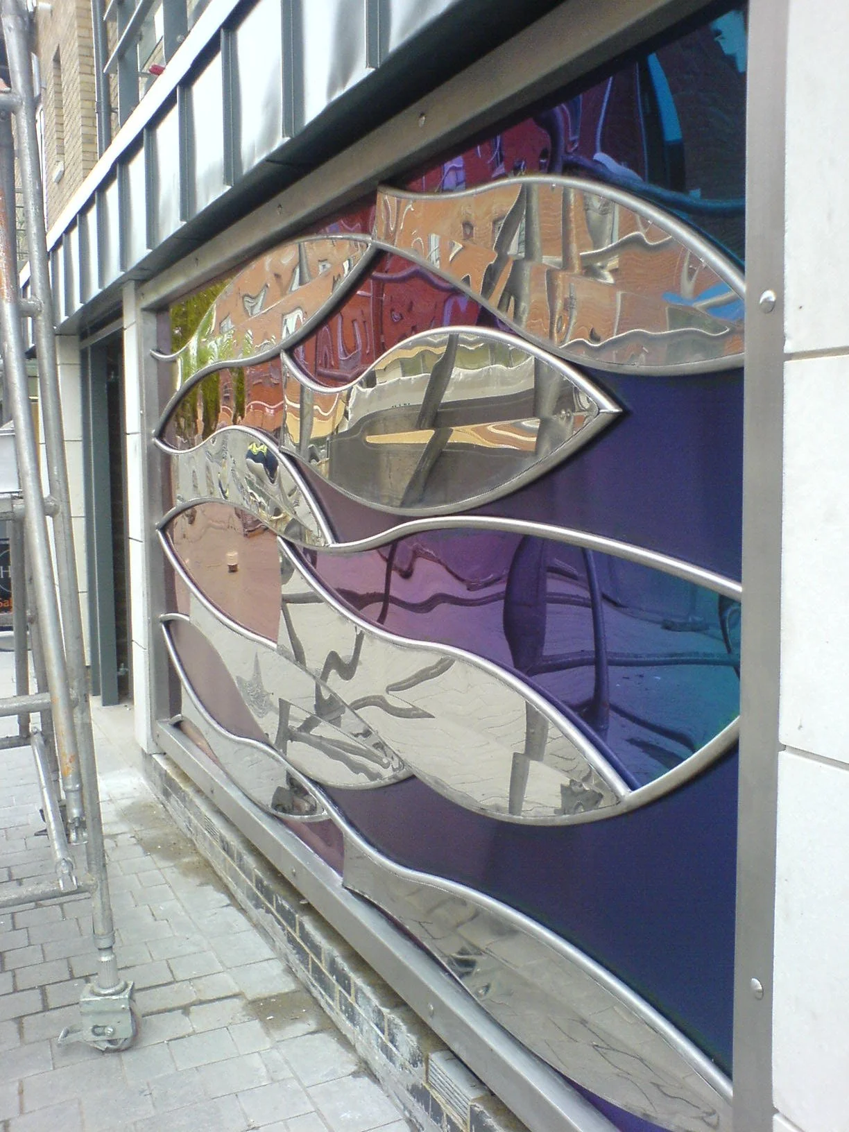 Decorative stained glass panel with abstract leaf design, mounted on a metal frame on an outdoor wall.