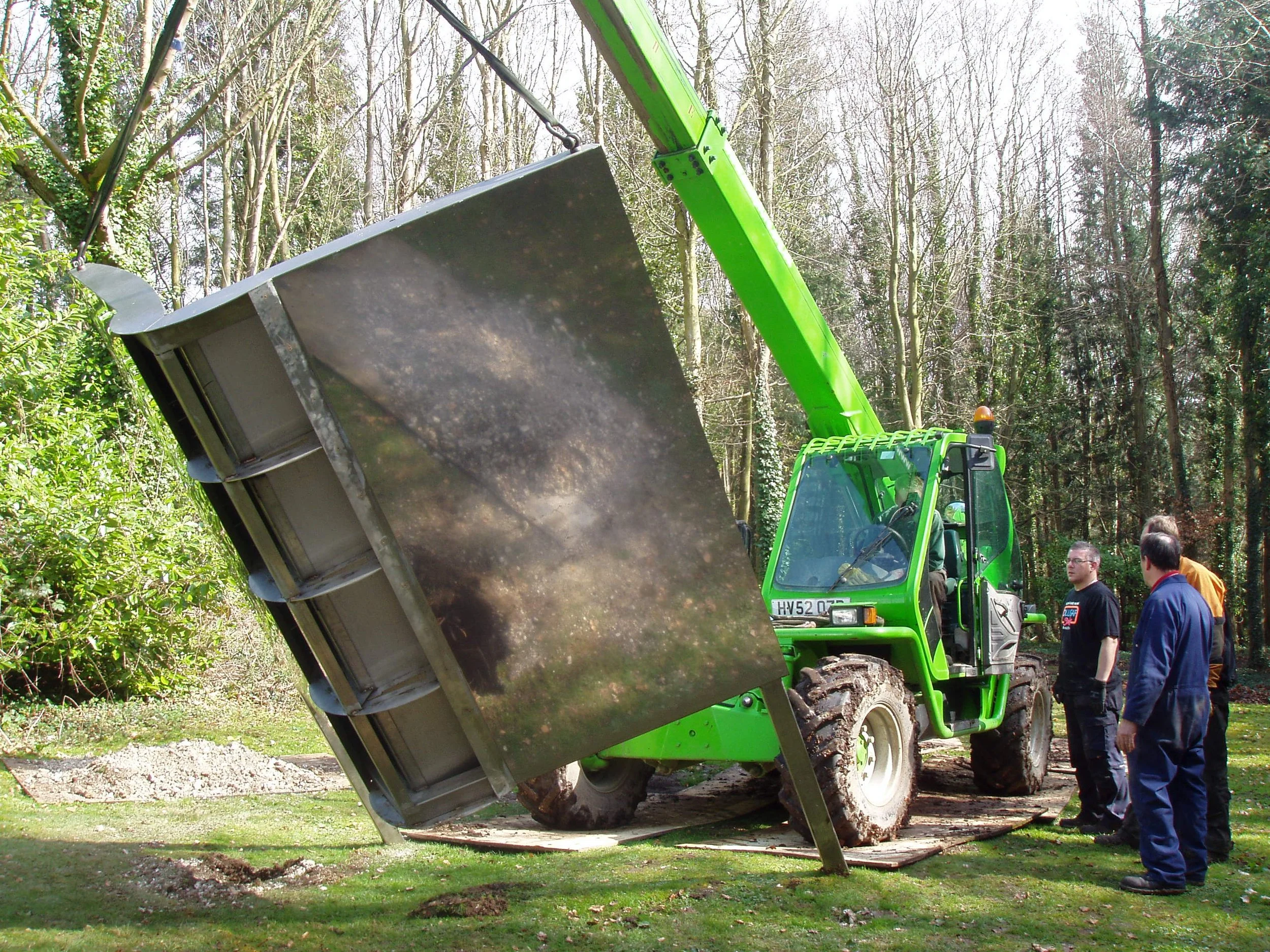 A large green construction vehicle with a tilted metal scoop lifting dirt or debris in a wooded area, with four men observing nearby.