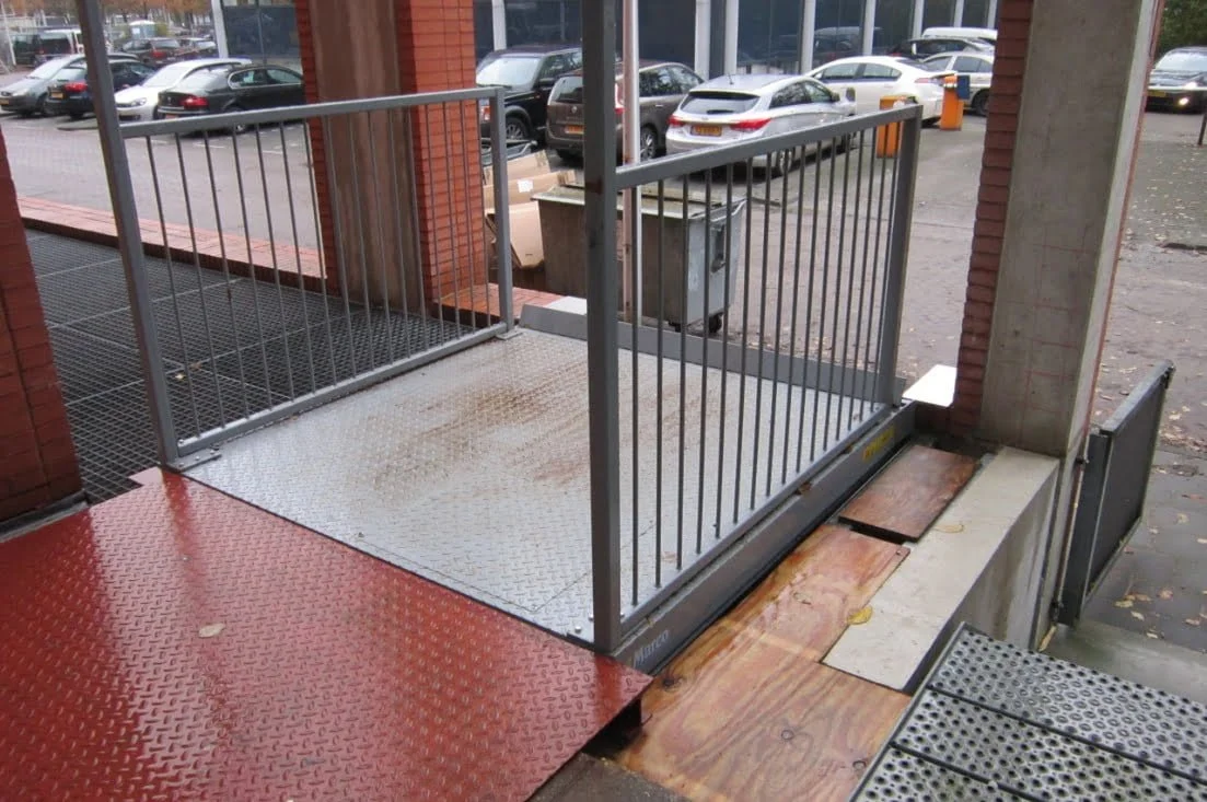 Image shows an outdoor wheelchair-accessible ramp with a metal railing, leading to a building entrance, with cars parked outside in the background.
