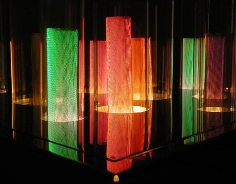Colorful illuminated cylindrical light fixtures in various colors including green, orange, and purple, arranged on a reflective surface in a dark setting.