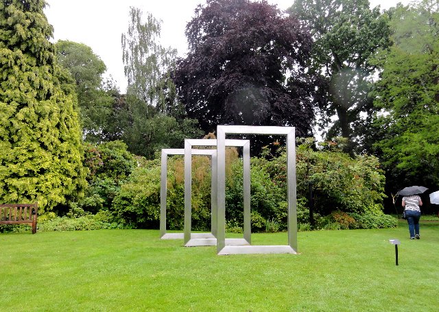 Modern outdoor art installation with three large white rectangular frames standing on a green lawn, surrounded by trees and bushes, with a person holding an umbrella walking in the background.